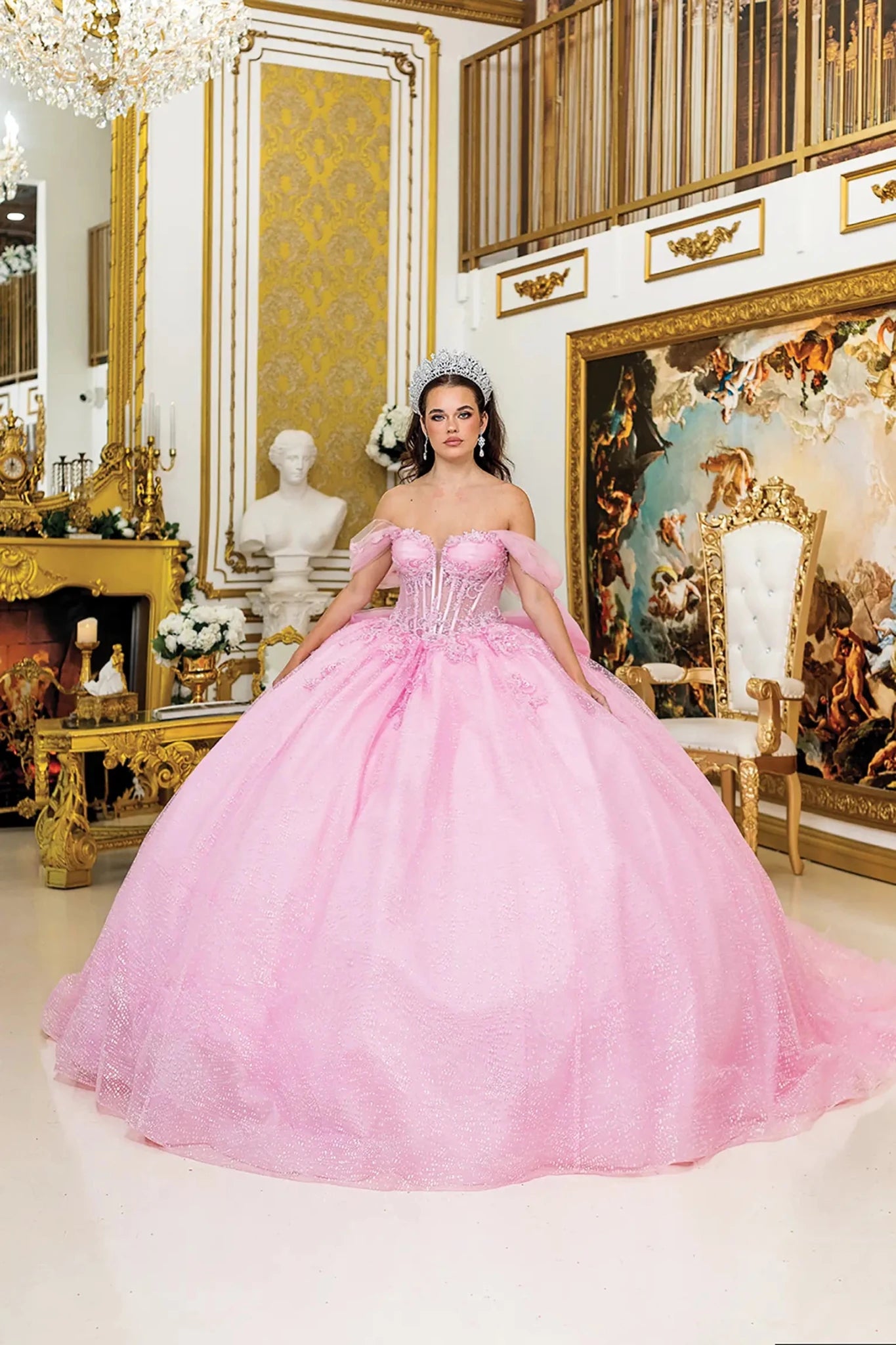 Woman standing front-facing in pink quinceañera ball gown with corset bodice, off-shoulder sheer puff sleeves, 3D floral appliqués, and voluminous glitter tulle skirt