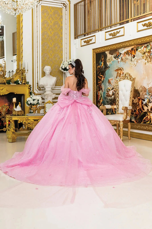 Woman standing back-facing in pink quinceañera ball gown with lace-up corset bodice, off-shoulder sheer puff sleeves, 3D floral appliqués, and flowing glitter tulle skirt with train