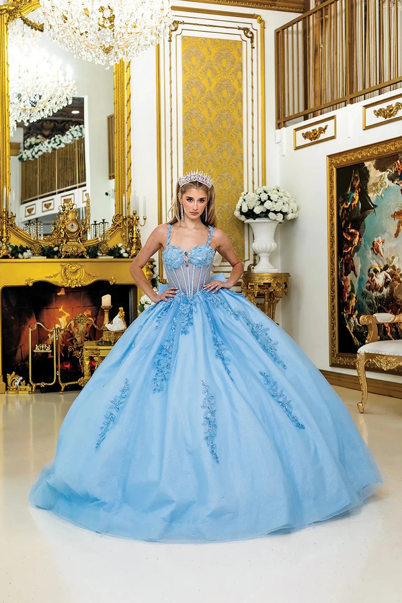 Front view of light blue quinceañera ball gown with sleeveless corset bodice, sheer boning detail, floral applique accents, and full glitter tulle skirt in an elegant interior