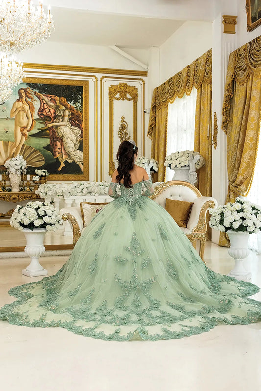 Woman standing back-facing in lilac-sage quinceañera ball gown with lace-up corset bodice, off-shoulder floral appliqués, glittered tulle skirt, and flowing layered train
