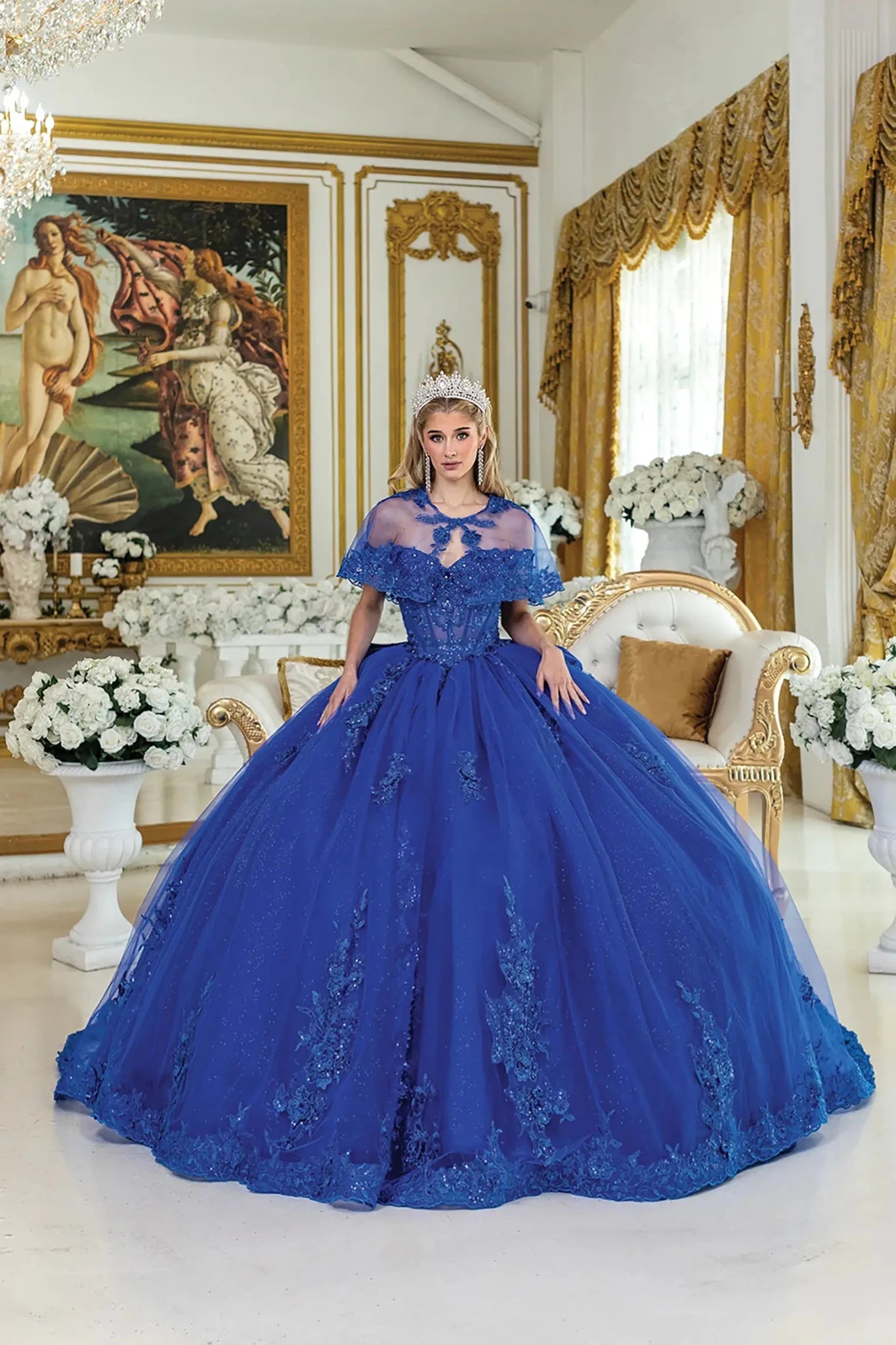 Woman standing front-facing in royal blue quinceañera ball gown with sheer capelet, corset bodice, lace appliqué detailing, beaded embellishments, and glitter tulle voluminous skirt