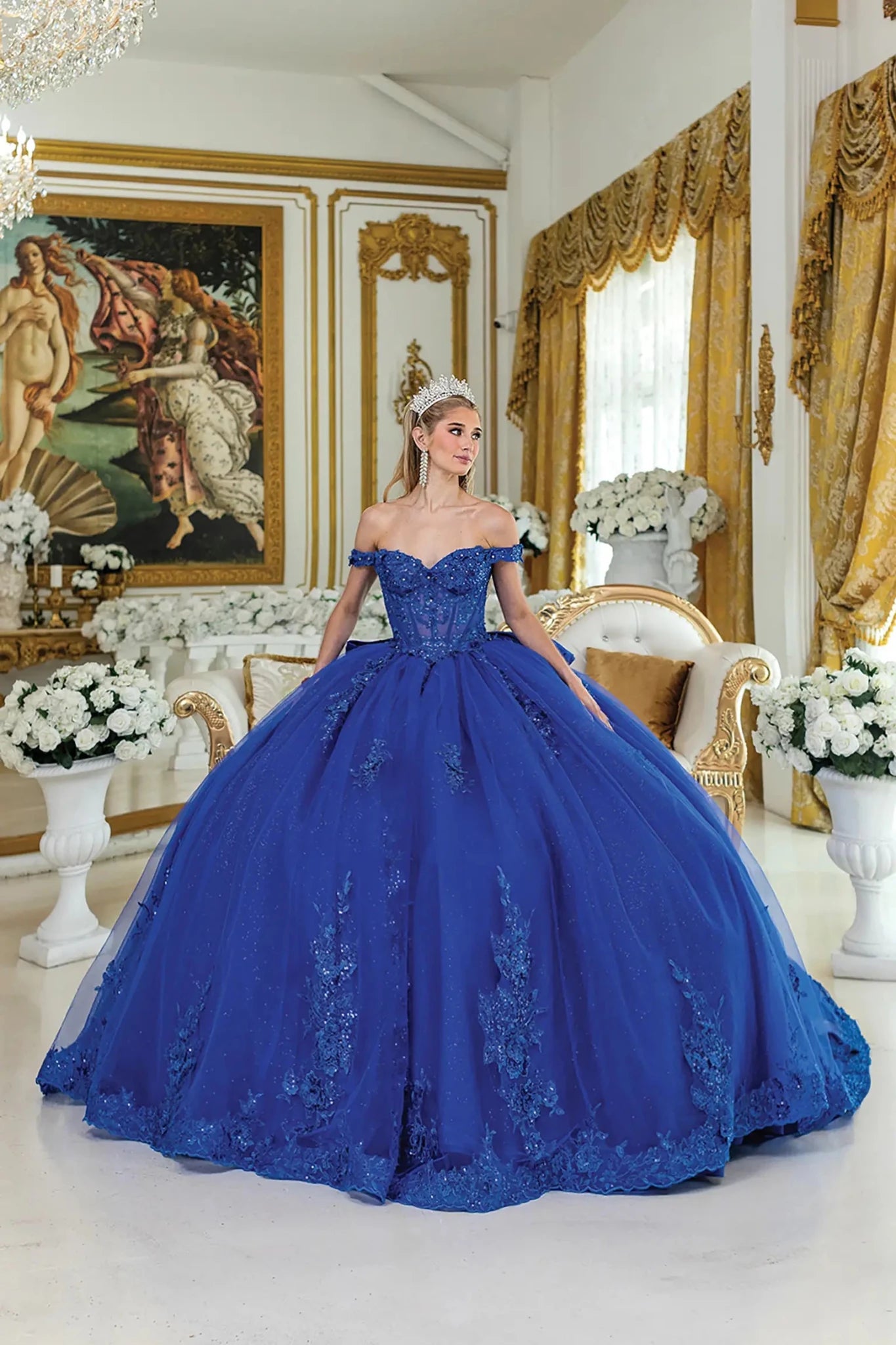 Woman standing front-facing in royal blue quinceañera ball gown with sheer capelet, corset bodice, lace appliqué detailing, beaded embellishments, and glitter tulle voluminous skirt