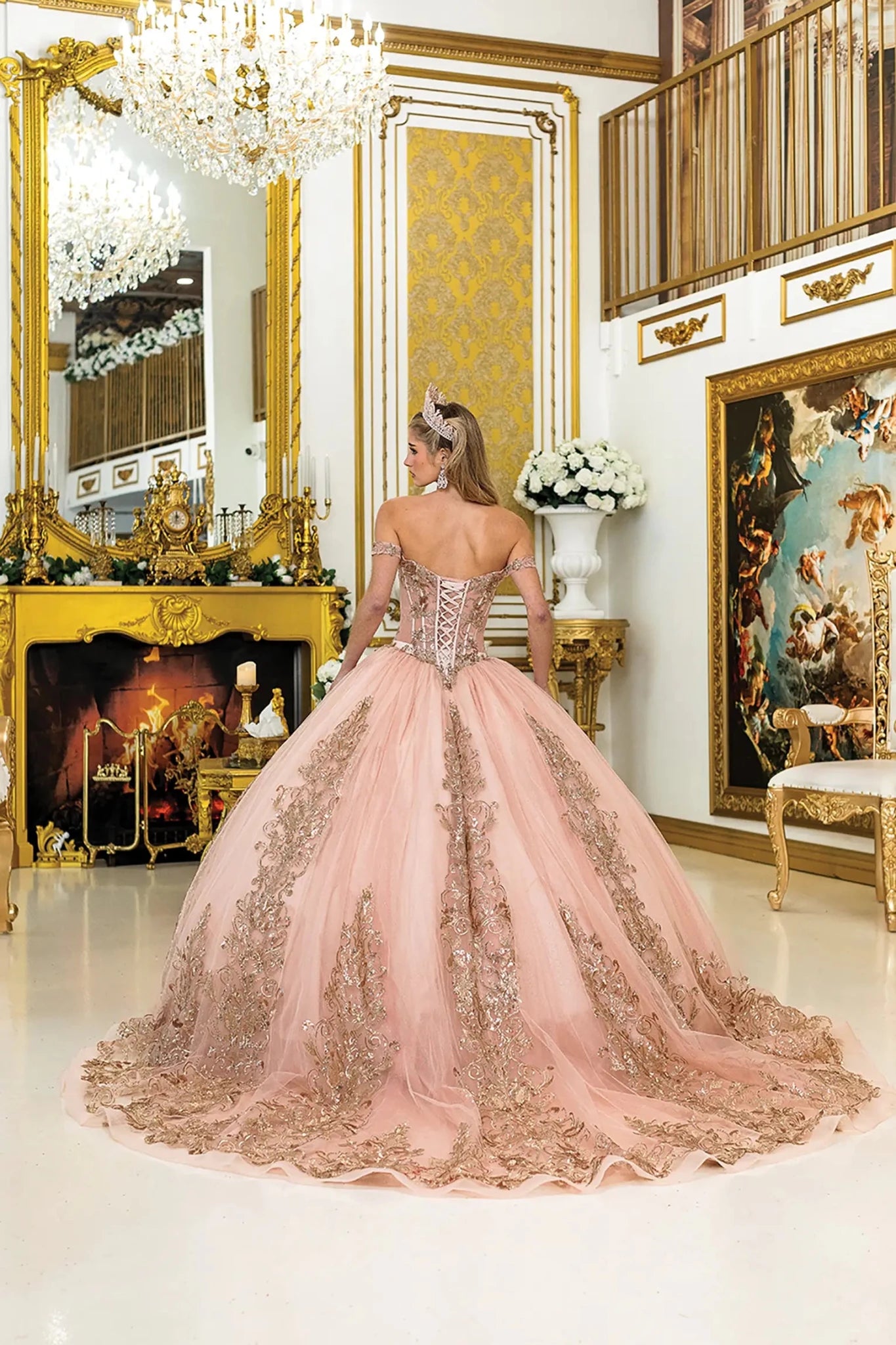 Back view of rose gold quinceañera ball gown with lace-up corset bodice, flowing tulle skirt adorned with lace appliqués, and a soft sweep train in a grand ballroom.