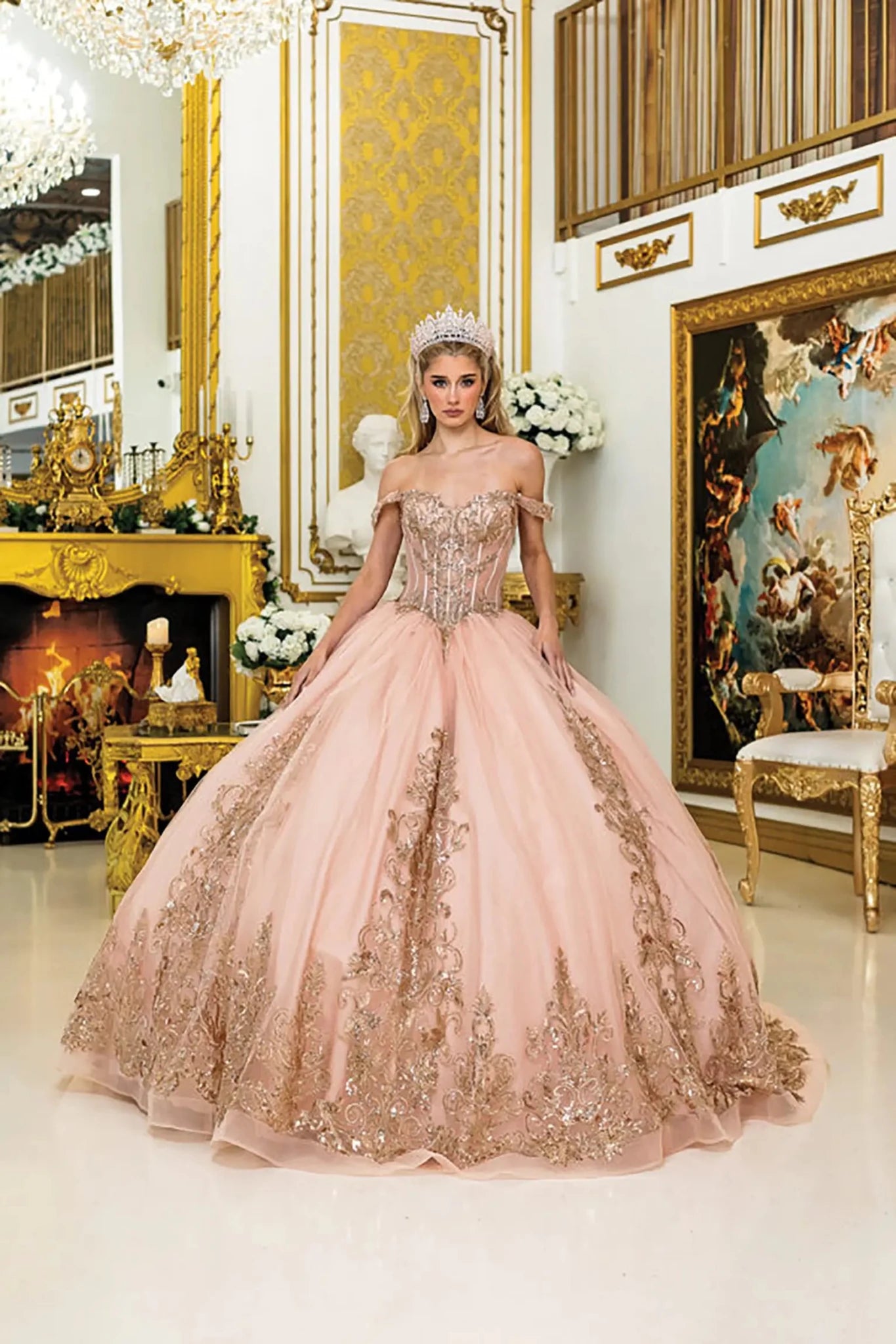 Woman standing front-facing in gold rose quinceañera ball gown with off-shoulder straps, corset bodice, tiered tulle skirt, intricate lace appliqués, and sparkling beaded embellishments