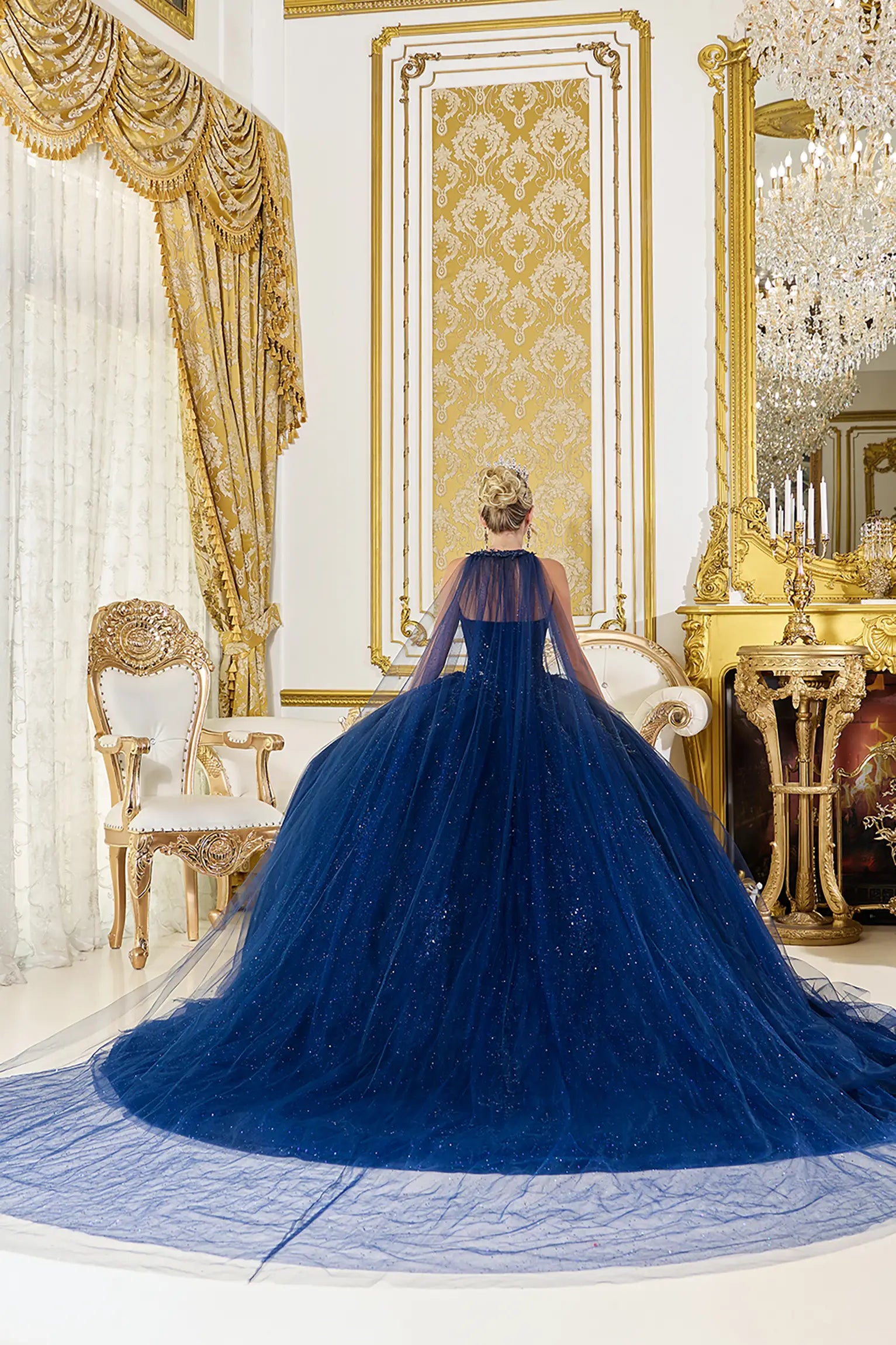 Back view: Navy blue quinceañera ball gown back view featuring a sheer cape-style overlay, flowing tulle skirt with subtle sparkle, and an extended train in an elegant ballroom interior
