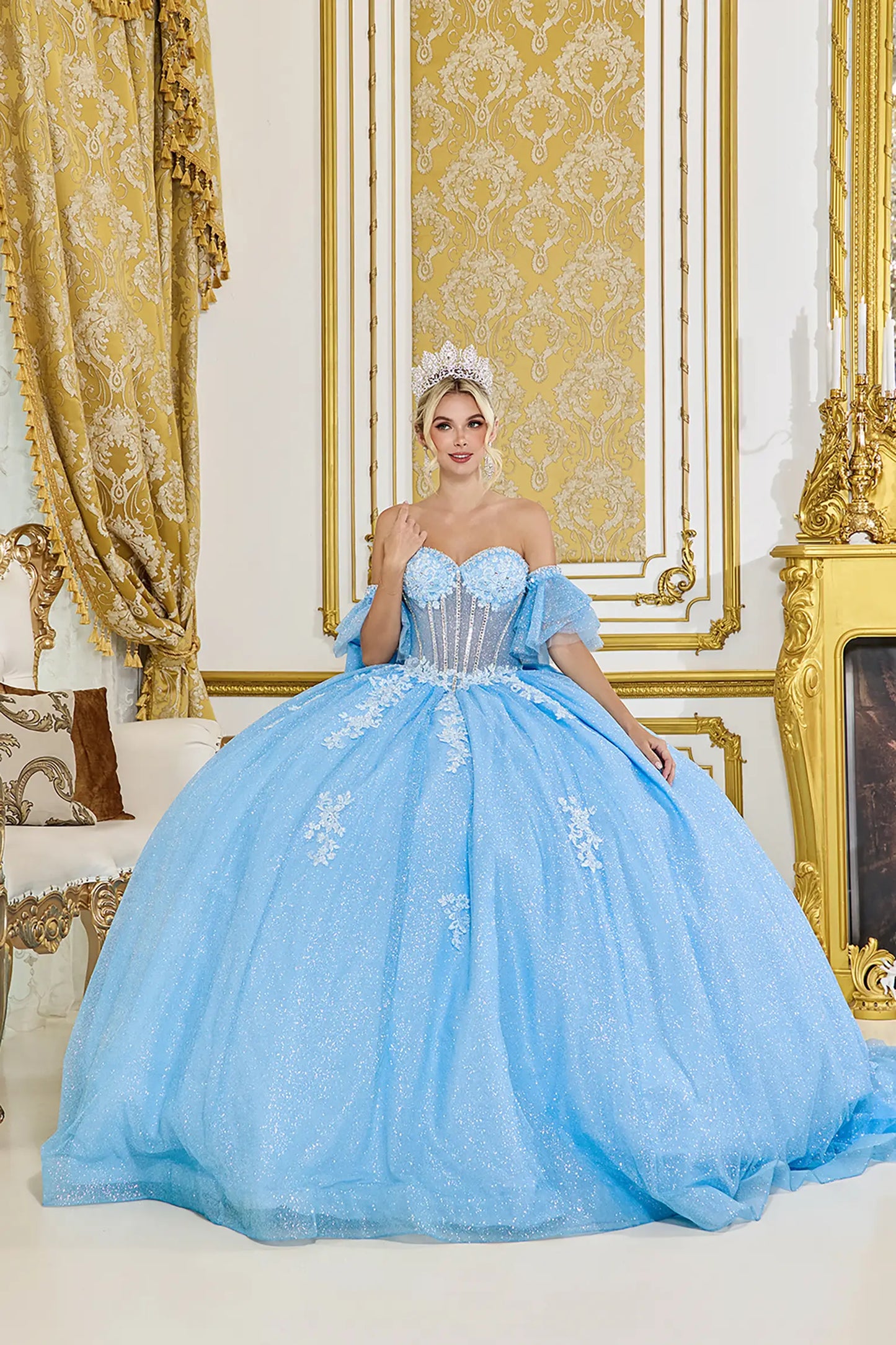 Front view: Light blue quinceañera ball gown with sweetheart corset bodice, off-the-shoulder tulle sleeves, floral lace appliqués, and a full glitter tulle skirt in an elegant palace setting