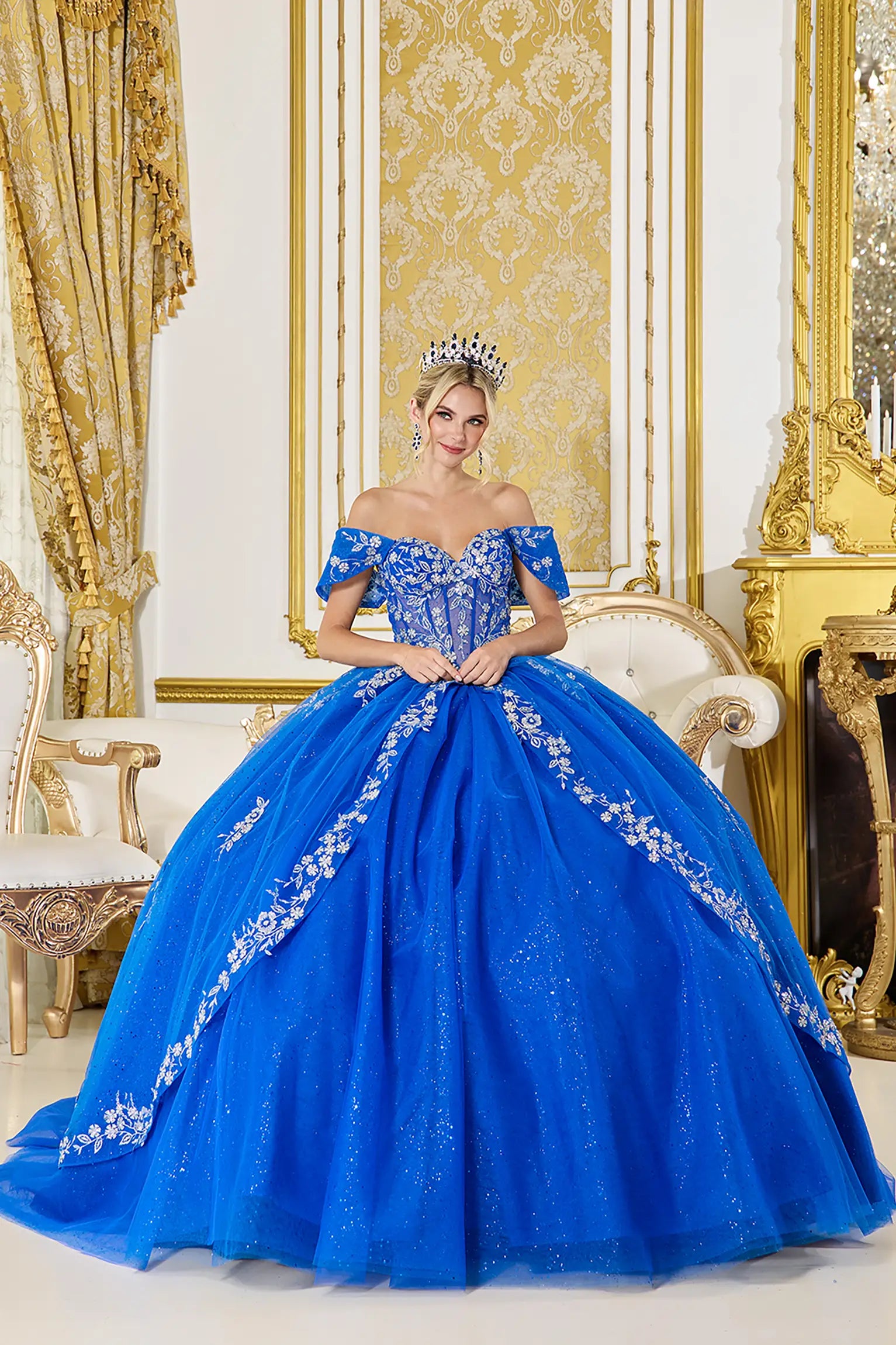 Front view of royal blue quinceañera ball gown featuring off-shoulder corset bodice, floral embroidery, glitter tulle skirt, and elegant tiara styling in a palace setting