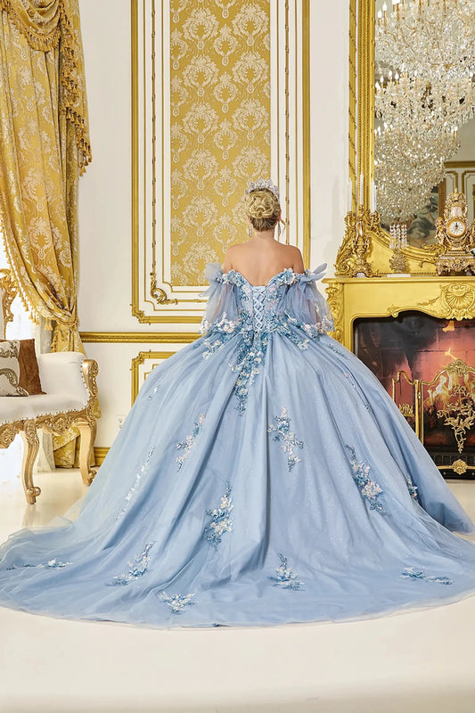 Back view of dusty blue quinceañera ball gown featuring lace-up corset back, sheer off-shoulder sleeves, floral appliqués, and flowing glitter tulle train in a luxurious setting