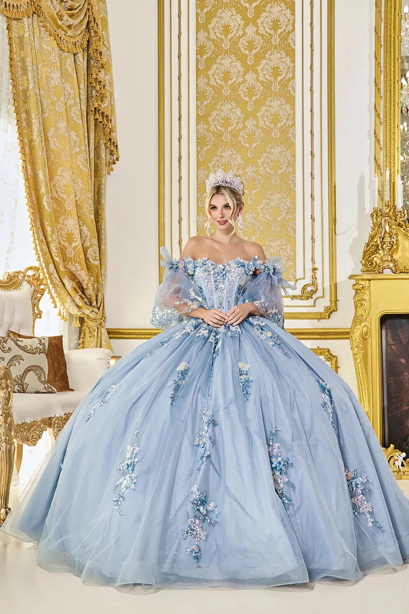 Front view of dusty blue quinceañera ball gown with off-shoulder sheer sleeves, corset bodice, floral appliqué detailing, and glitter tulle skirt styled in an elegant palace interior