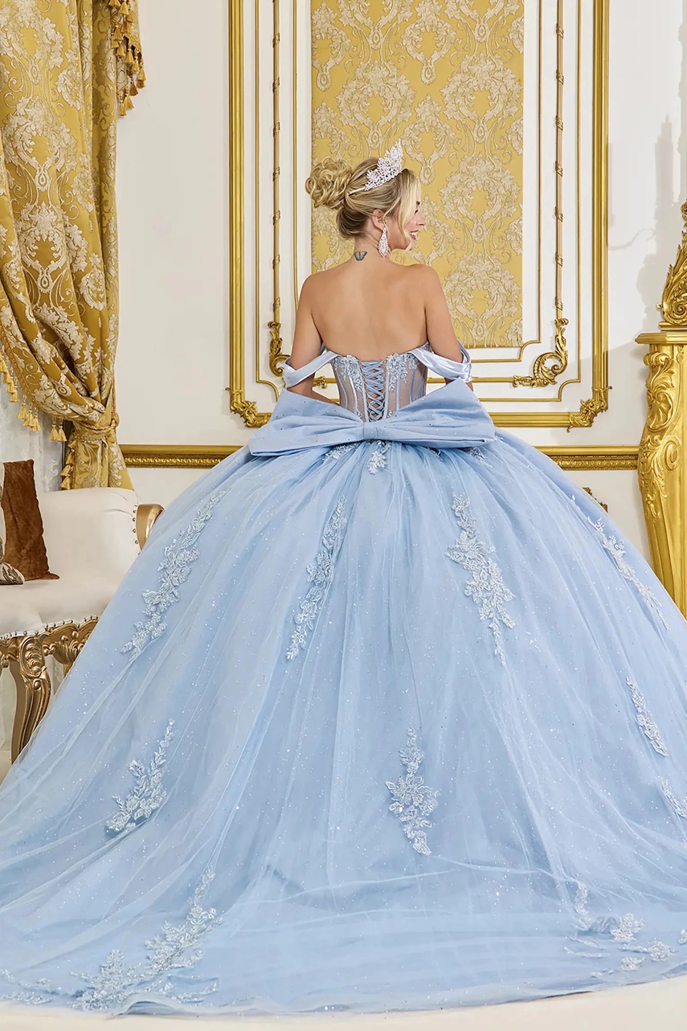 Back view of dusty blue quinceañera ball gown with off-the-shoulder straps, lace-up corset back, oversized satin bow, glitter tulle skirt, and floral lace appliqués
