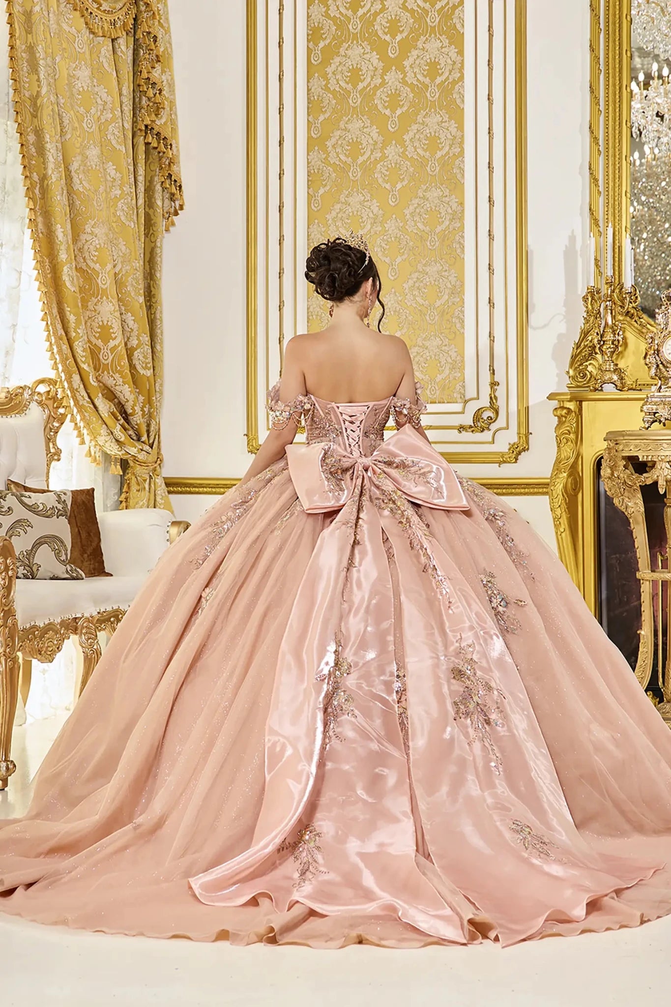 Back view of rose gold quinceañera ball gown featuring a lace-up corset back, oversized satin bow, and long flowing train with delicate applique details