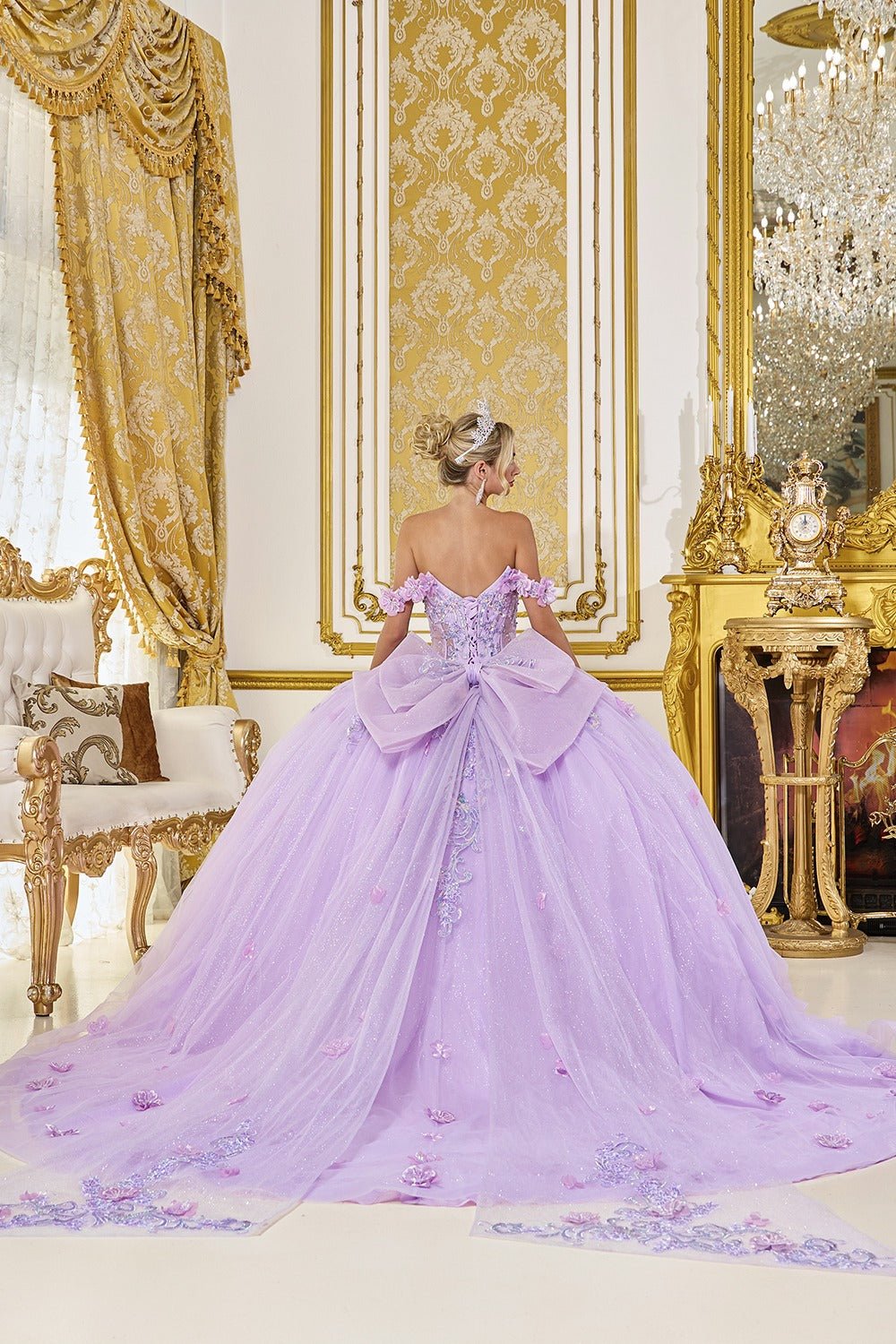 Woman in a lilac ball gown with a big back ribbon and train standing in an opulent room with gold decor.