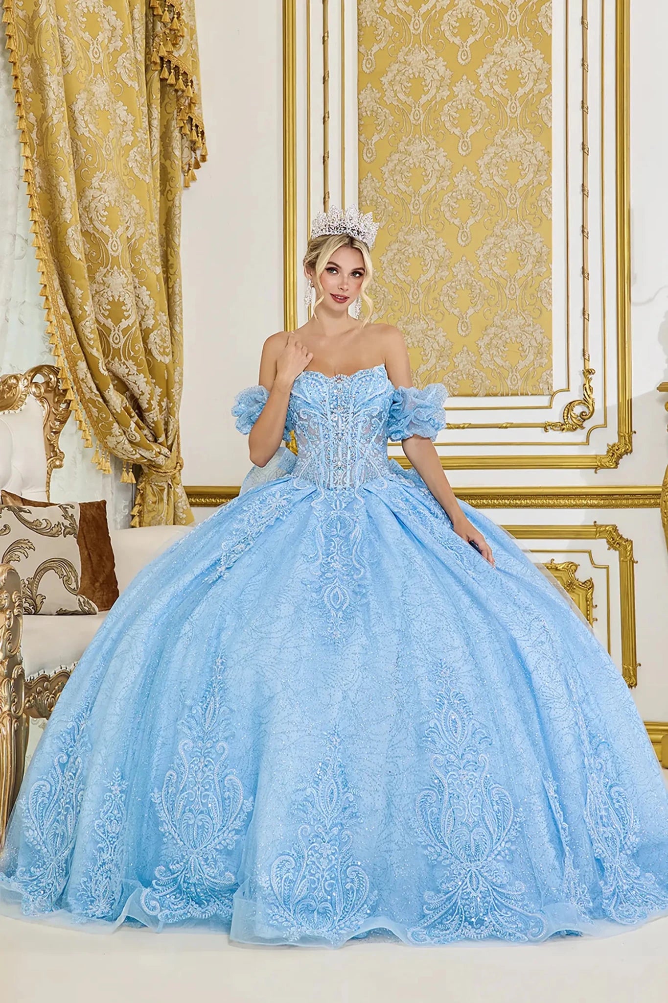 Front view of light blue quinceañera ball gown with off-the-shoulder ruffled sleeves, embellished corset bodice, and full glitter lace skirt in an elegant interior