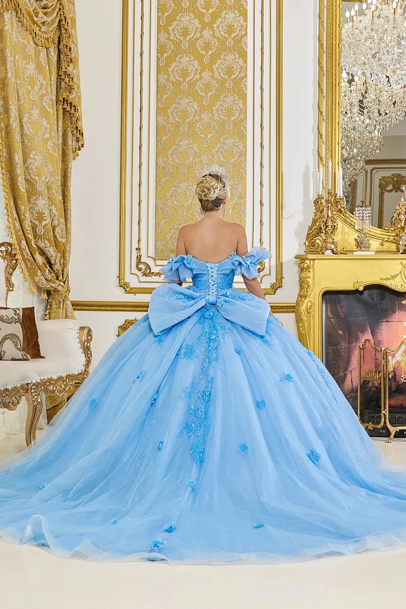 Woman standing back-facing in Bahama blue ball gown with lace-up closure, 3D floral appliqué accents, ruffle sleeves, and cascading skirt