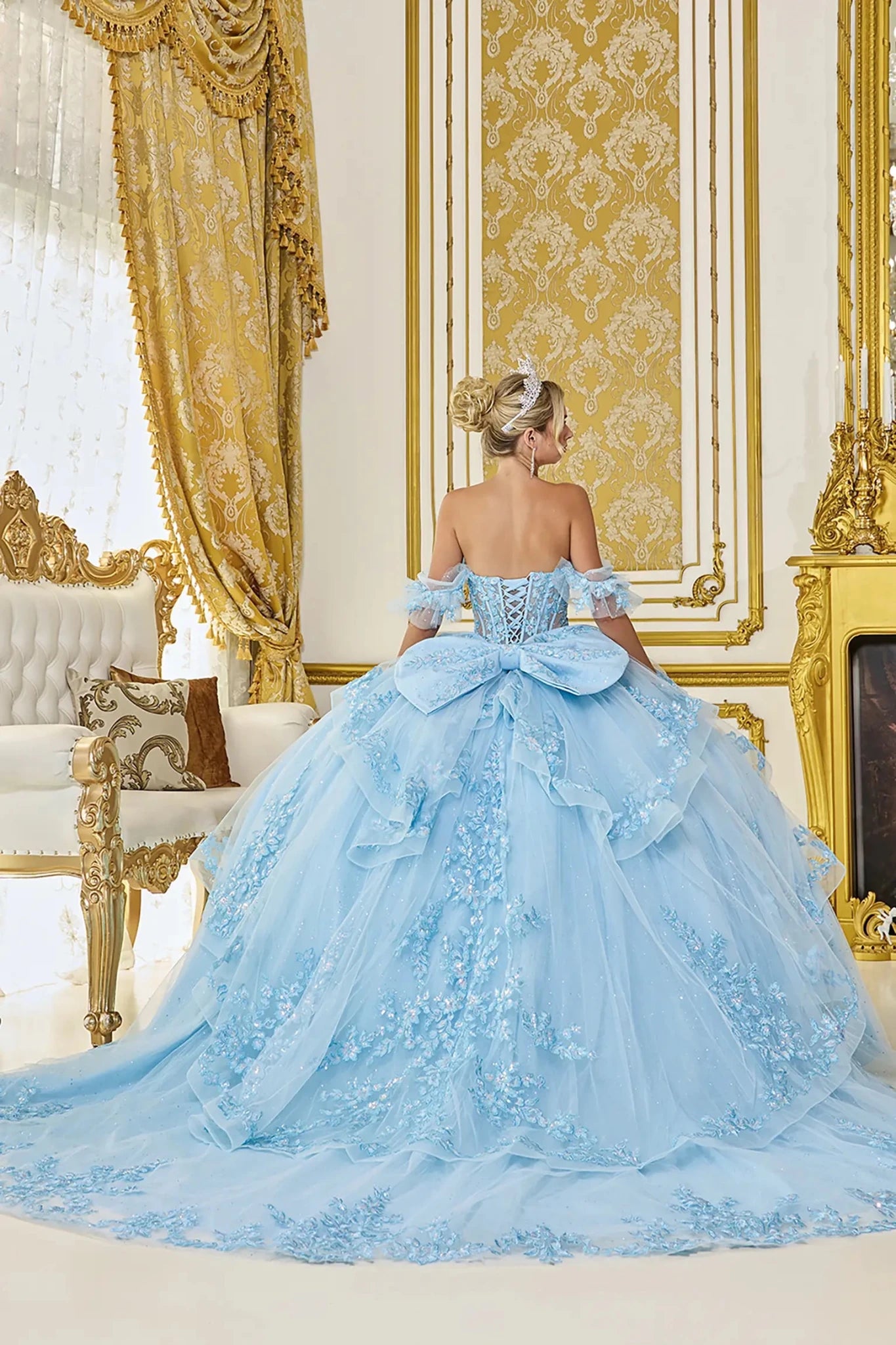 Back view of light blue quinceañera ball gown featuring a lace-up corset back, oversized bow detail, and long flowing tulle train with delicate applique accents