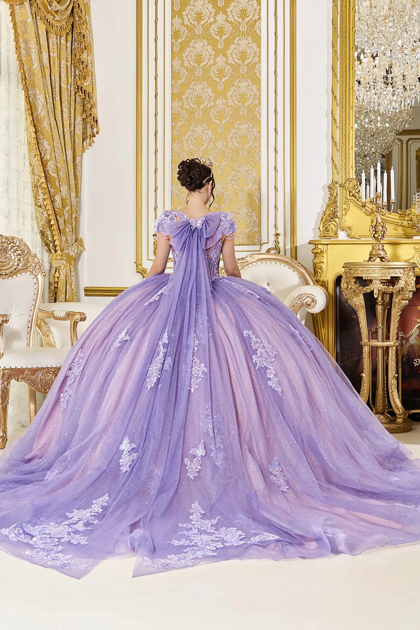 Back view of lilac quinceañera ball gown featuring a soft bow with flowing tulle tails, lace-up corset back, cap sleeves, and voluminous glitter tulle skirt with floral appliqués.