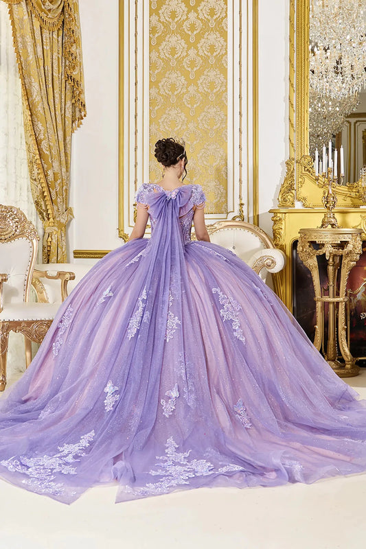Back view of lilac quinceañera ball gown featuring a soft bow with flowing tulle tails, lace-up corset back, cap sleeves, and voluminous glitter tulle skirt with floral appliqués.