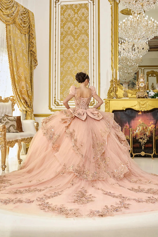 Woman standing back-facing in rose gold quinceañera ball gown with lace-up bodice, detachable cap sleeves, glitter floral appliqués, and flowing ruffled tiered skirt with train