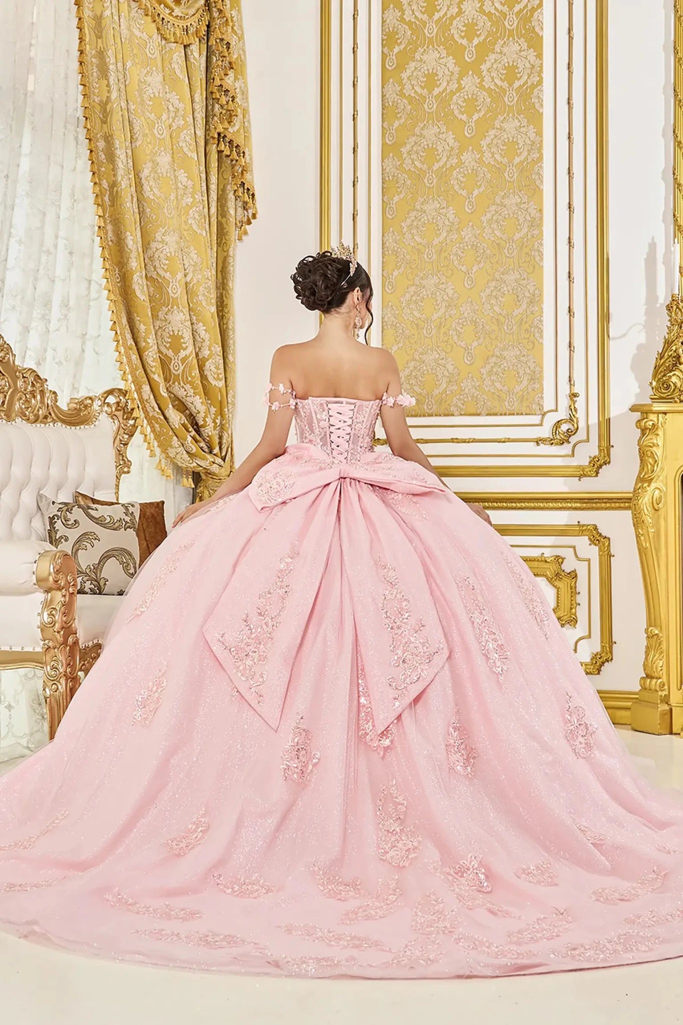 Woman back-facing in blush off-shoulder sweetheart quinceañera dress with lace-up corset bodice, embroidery, glitter tulle, and cascading full skirt