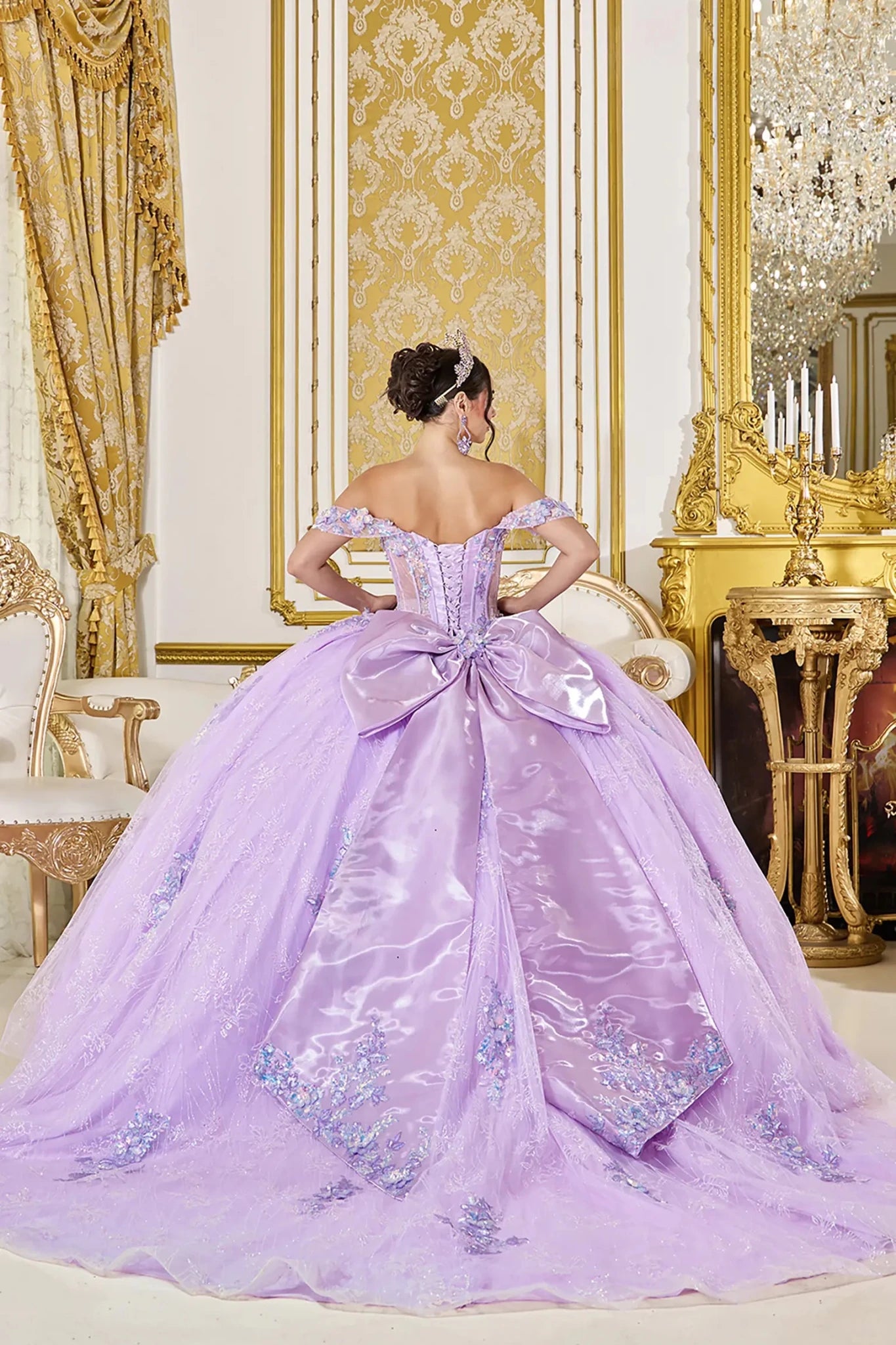 Back view of lilac quinceañera ball gown featuring a lace-up corset back, oversized satin bow, and long flowing tulle train with embellished applique accents