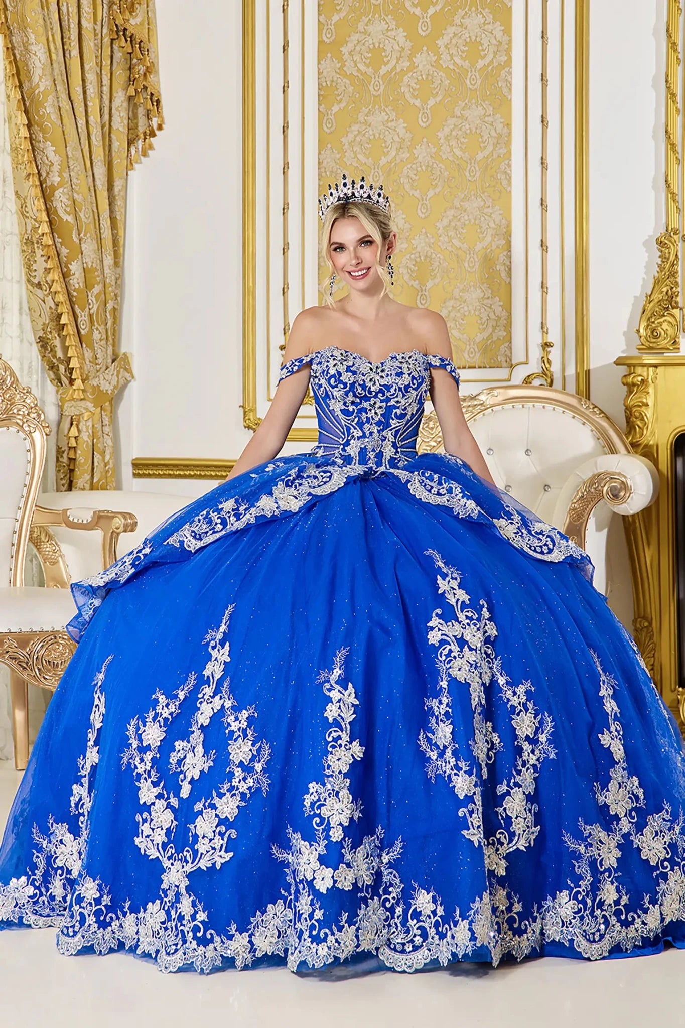 Front view of royal blue quinceañera ball gown with off-the-shoulder sweetheart bodice, intricate lace appliques, and full glitter tulle skirt in an ornate interior