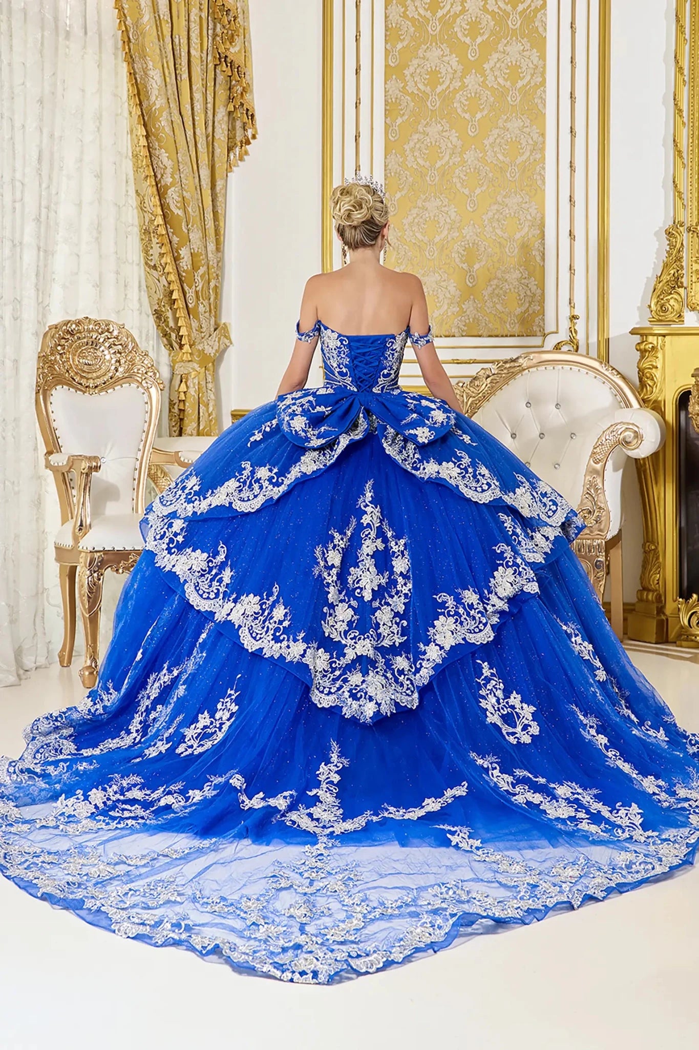 Back view of royal blue quinceañera ball gown featuring a lace-up corset back, layered overskirt design, and long flowing train with ornate lace applique detailing