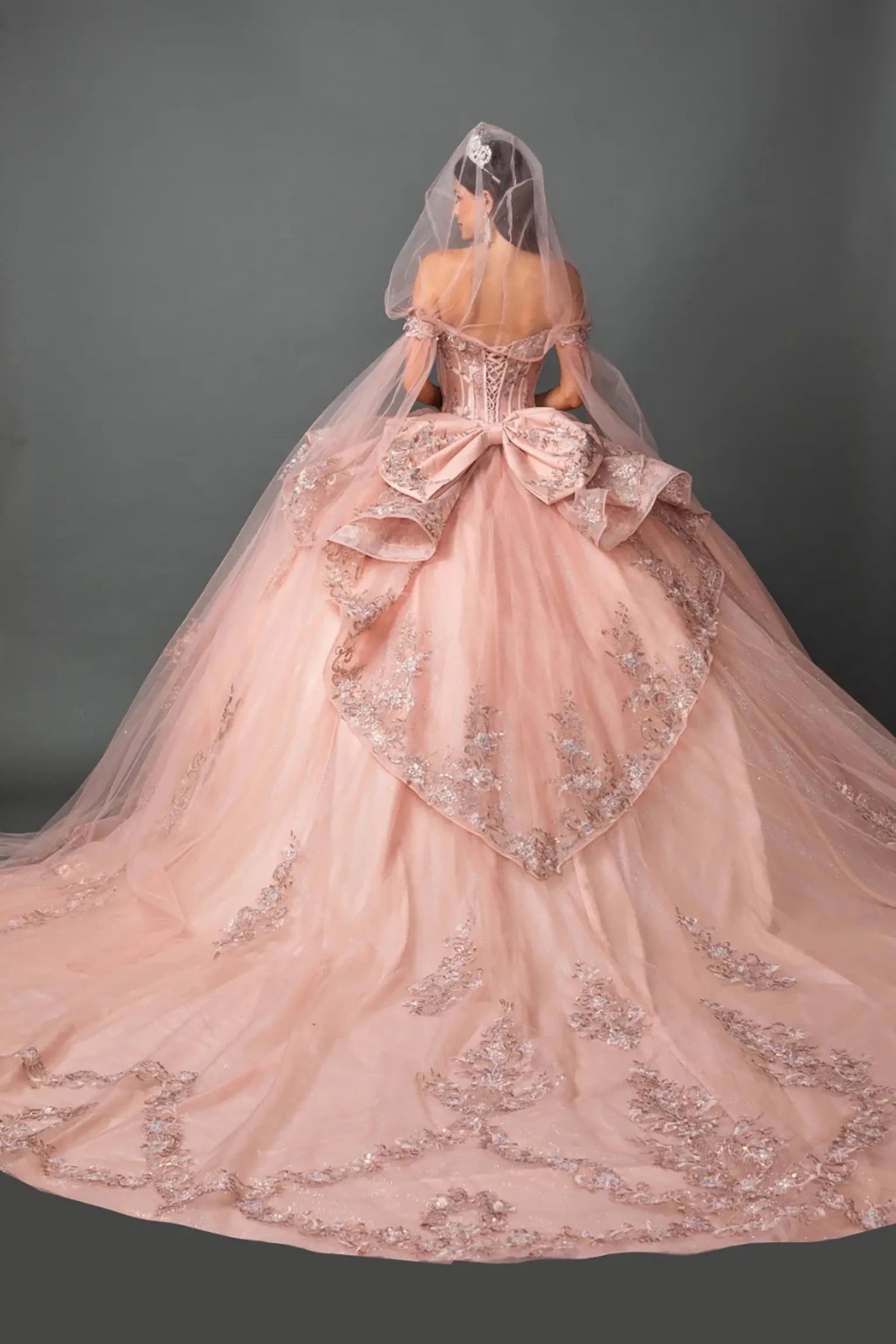 Woman standing back-facing in rose gold ball gown with lace-up closure, floral appliqué accents, glitter tulle layers, and cascading skirt
