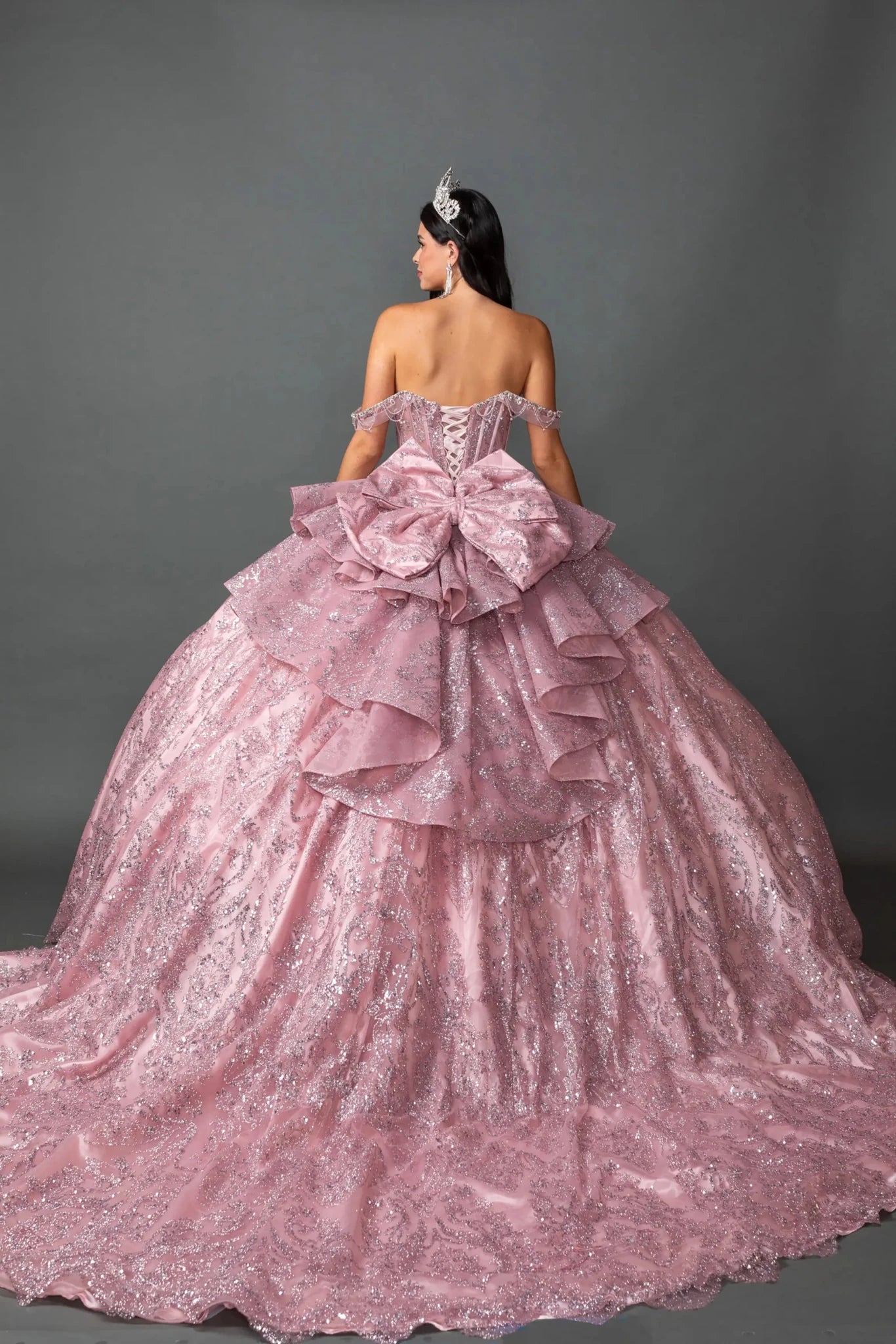 Woman standing back-facing in dusty pink off the shoulder sweetheart quinceañera ball gown with lace-up beaded corset bodice and glitter embroidered layered skirt with flowing train