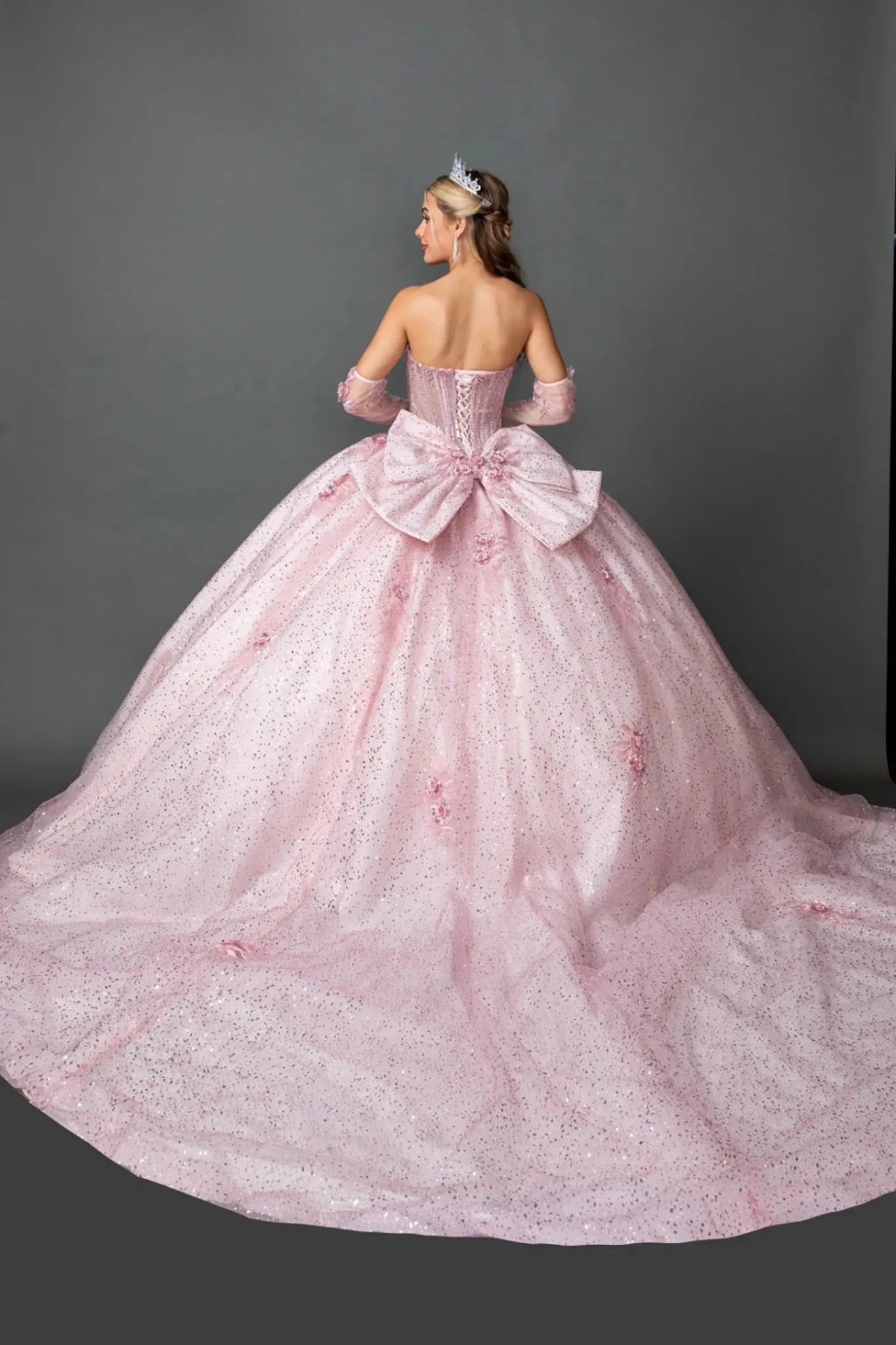 Back view of a woman wearing blush quinceañera, lace-up corset, glitter tulle, floral appliques, removable gloves
