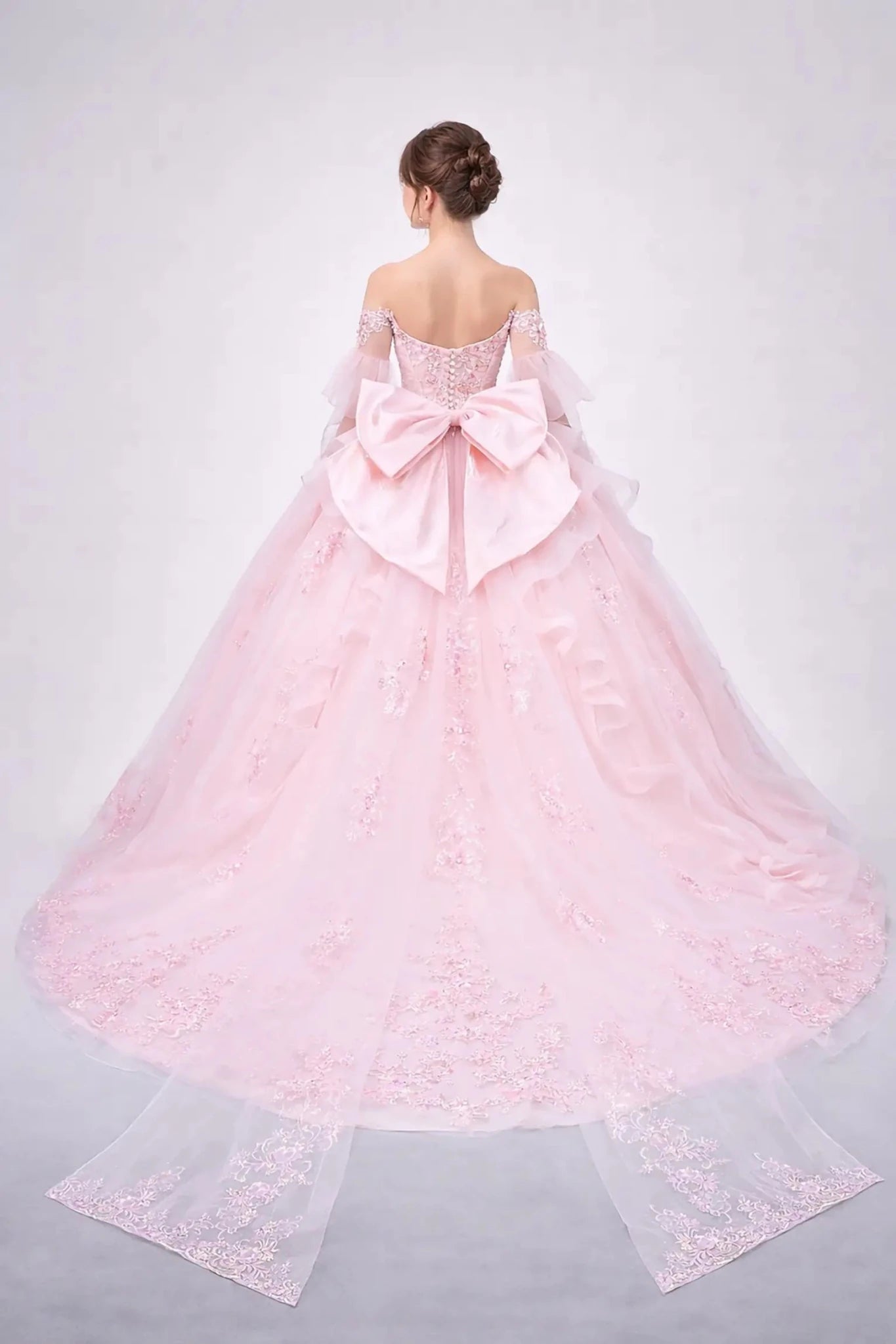 Woman standing back-facing in blush ball gown with lace-up corset bodice, embellished detailing, and cascading full skirt

