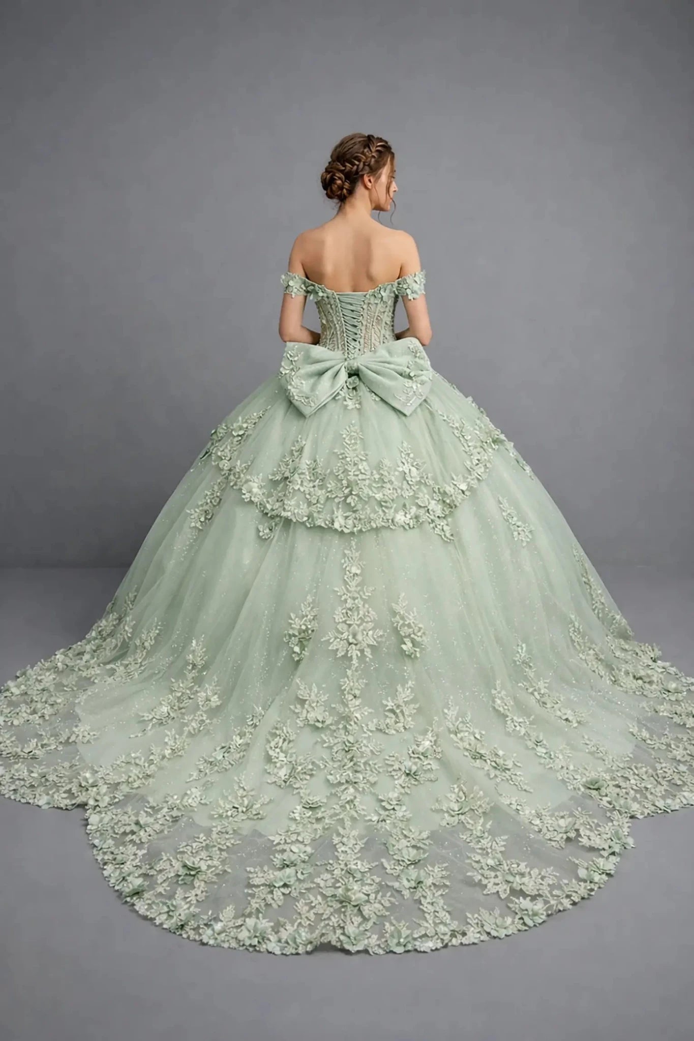 Woman standing back-facing in sage ball gown with lace-up closure, 3D floral appliqué accents, ruffle sleeves, and cascading skirt