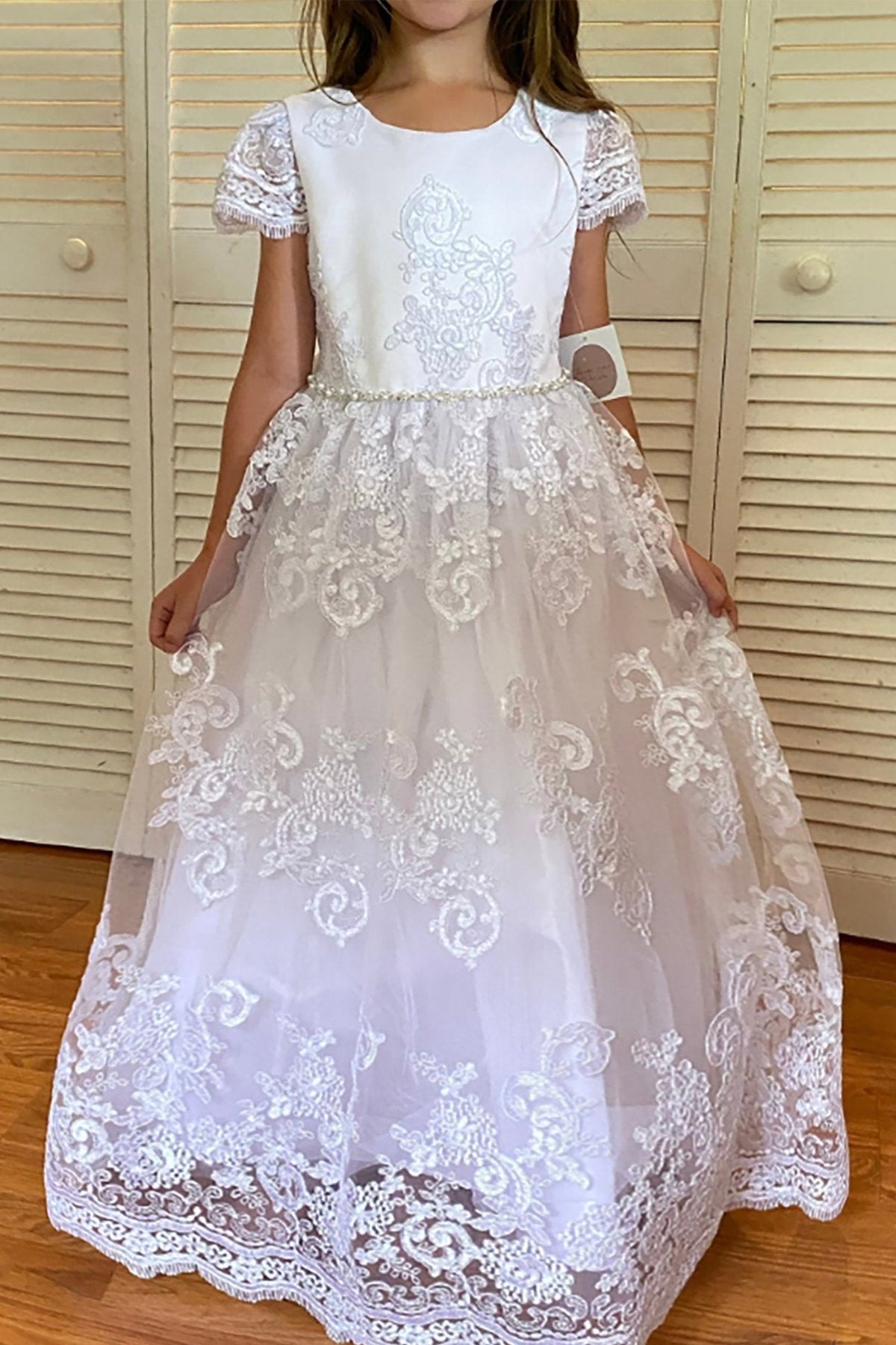 Detailed view highlights white full-length communion dress with lace cap sleeves, floral lace appliques, and pearl trim at the waist.