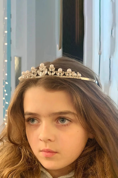 Young girl wearing a rhinestone and pearl flower motif silver tiara with a light blue curtain background