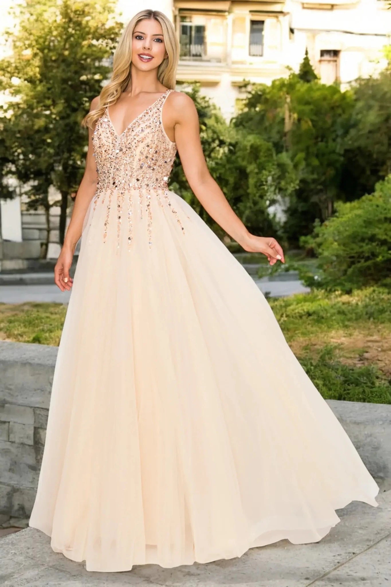 A full-length front view of a woman wearing a champagne formal dress with an open V-neckline and flowing skirt.