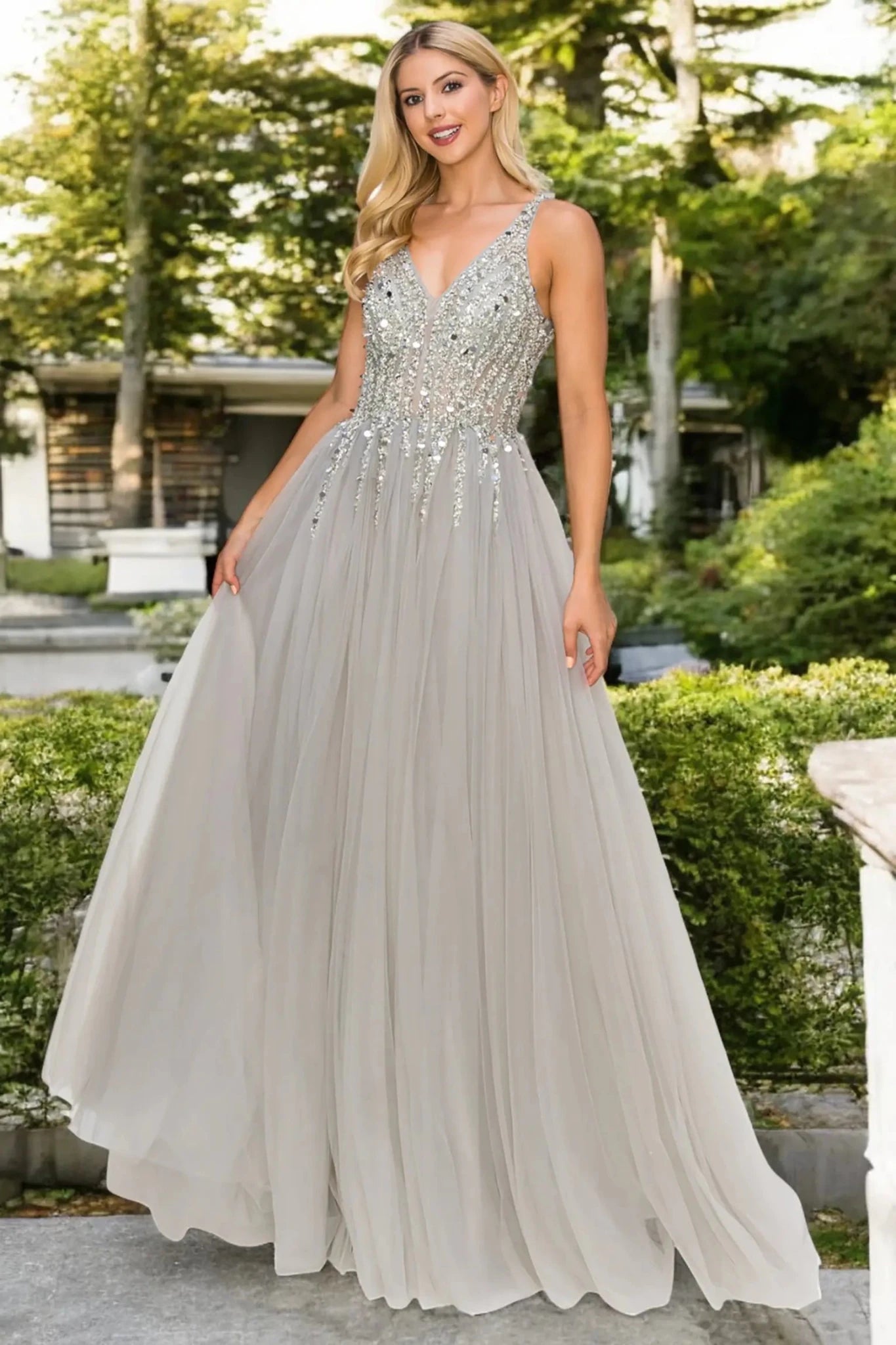 A full-length front view of a woman wearing a silver formal dress with an open V-neckline and flowing skirt.