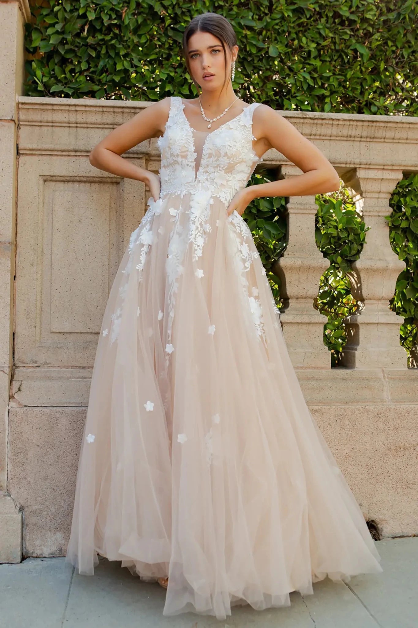 Full front view of a women wearing champagne floral lace A-line prom dress with illusion V-neck bodice and flowing skirt.