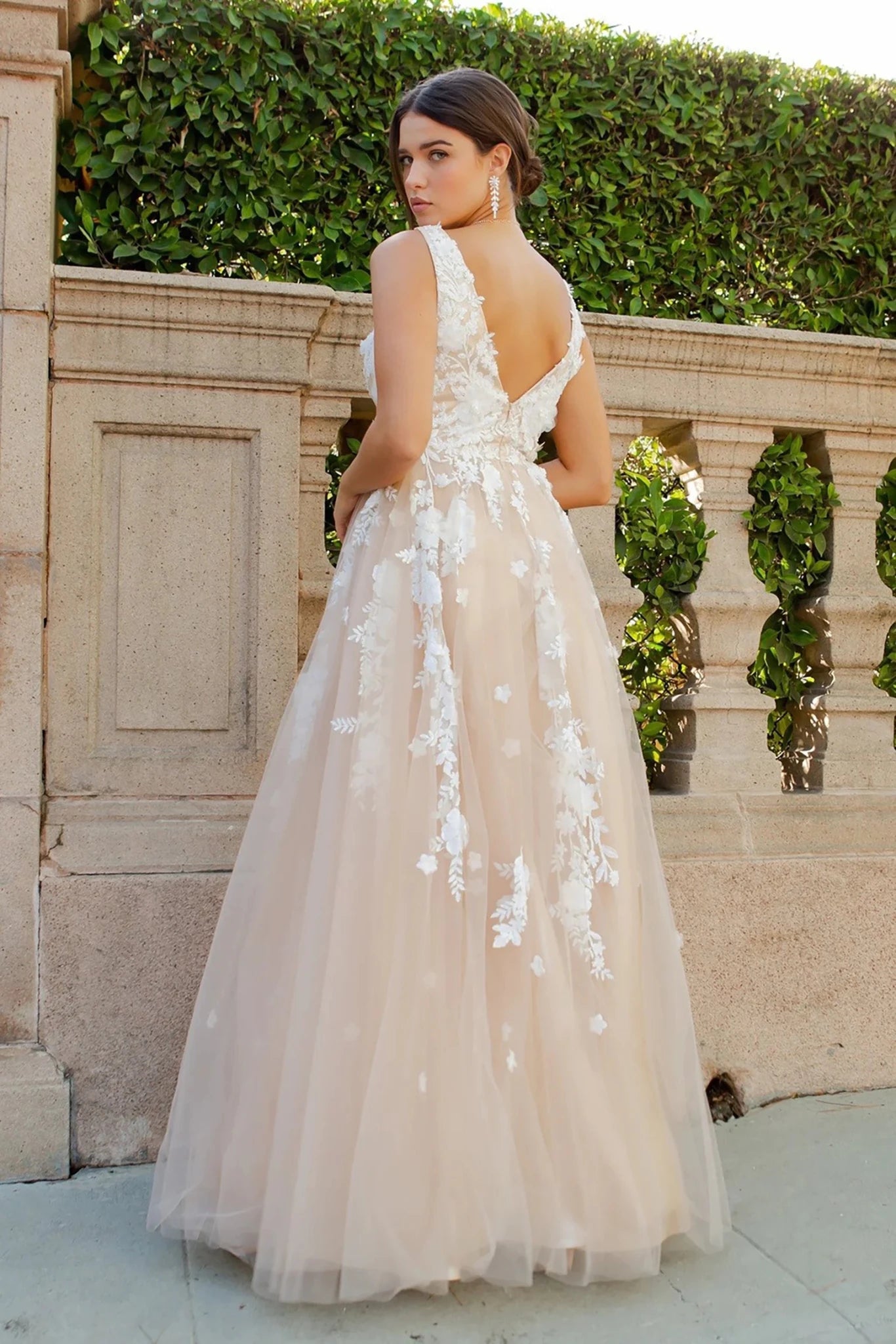 A woman wearing a sleeveless champagne tulle ball gown with white floral lace appliques and v back
