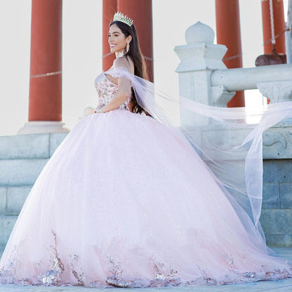 Displayed in a angled view, the Blush quinceañera dress has floral hem appliques and a sheer shoulder cape.
