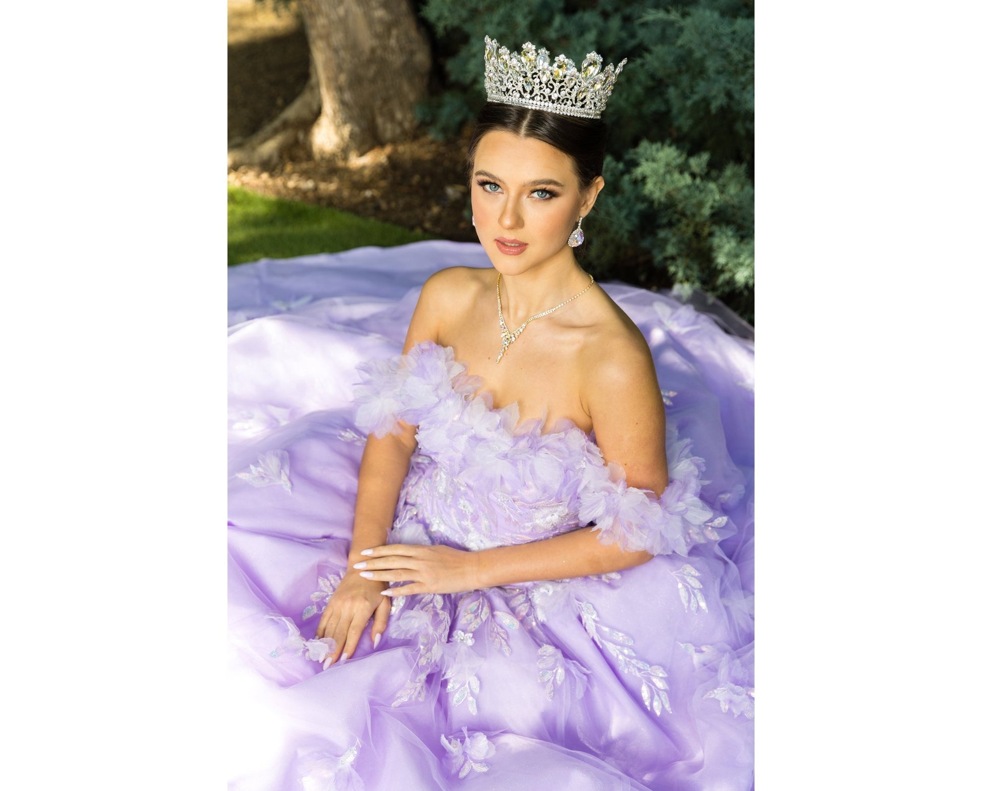 Sitting view of a woman wearing a lilac quinceanera dress with rainbow embroidery, sequin ruffle flowers, crown, and hands clasped in front