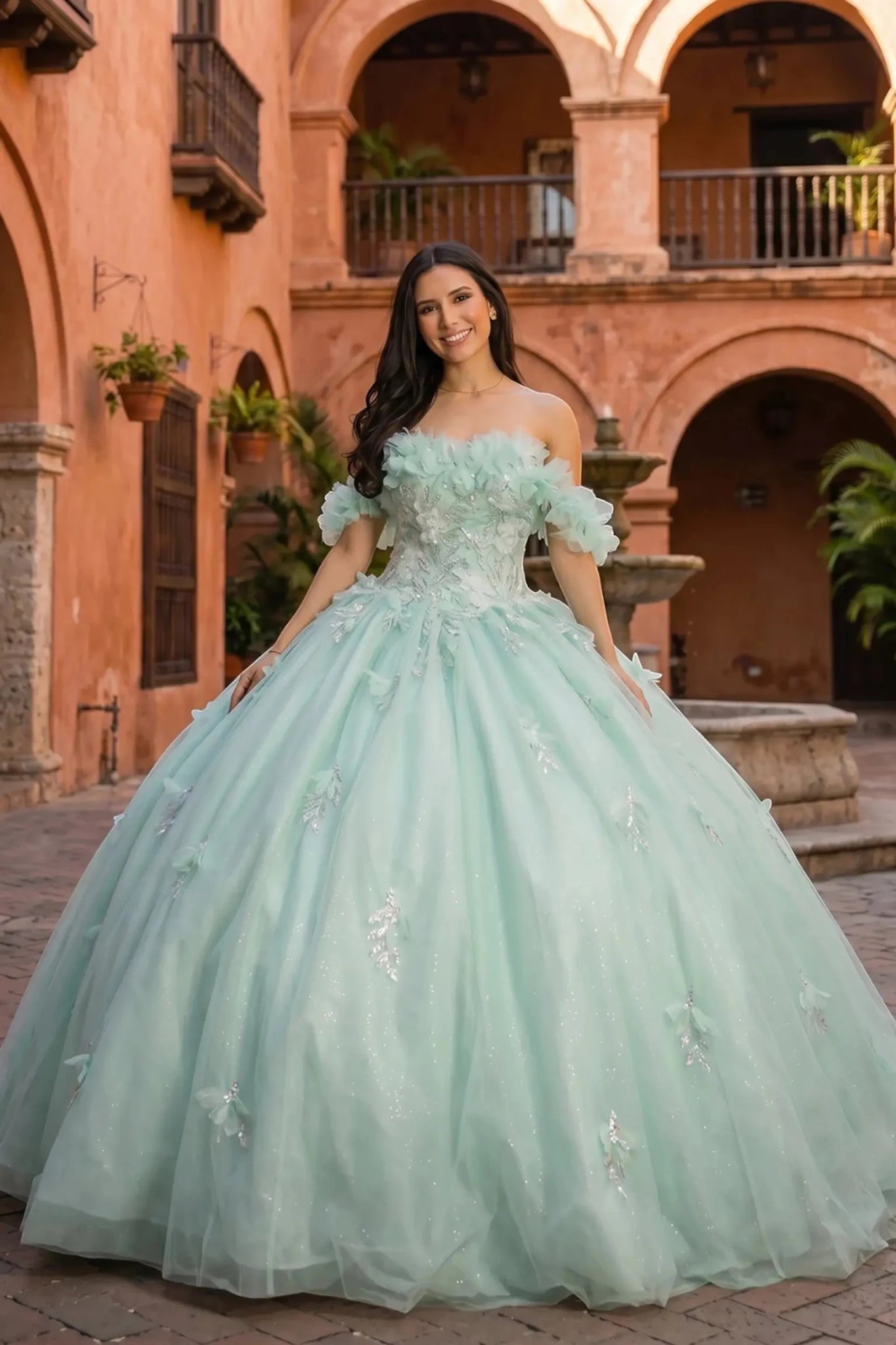 Front view of a woman wearing an aqua blue quinceanera dress with rainbow embroidery, sequin ruffle flowers, and off-shoulder neckline