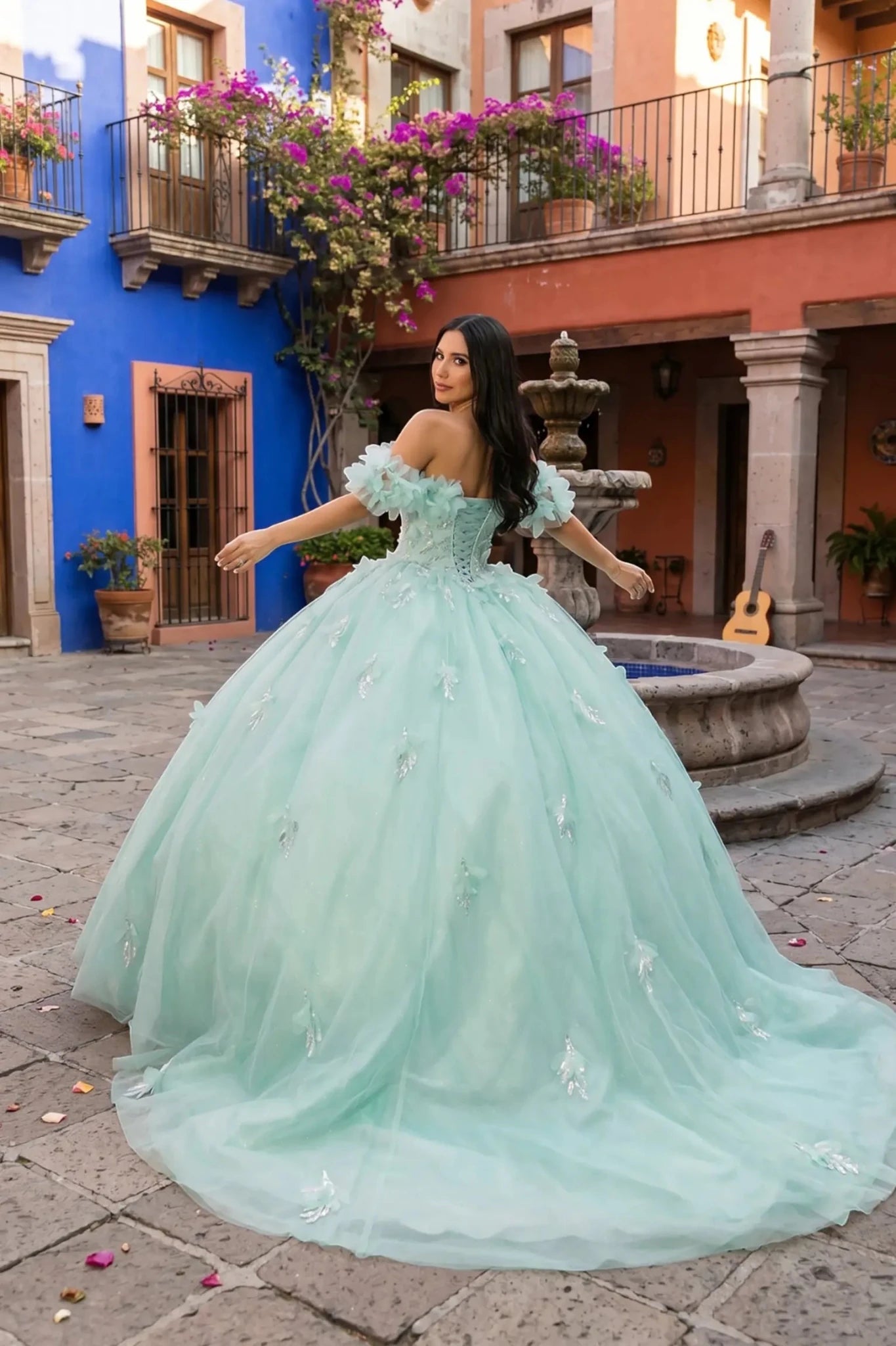 Back view of a woman wearing an aqua blue quinceanera dress with rainbow embroidery, sequin ruffle flowers, and off-shoulder neckline