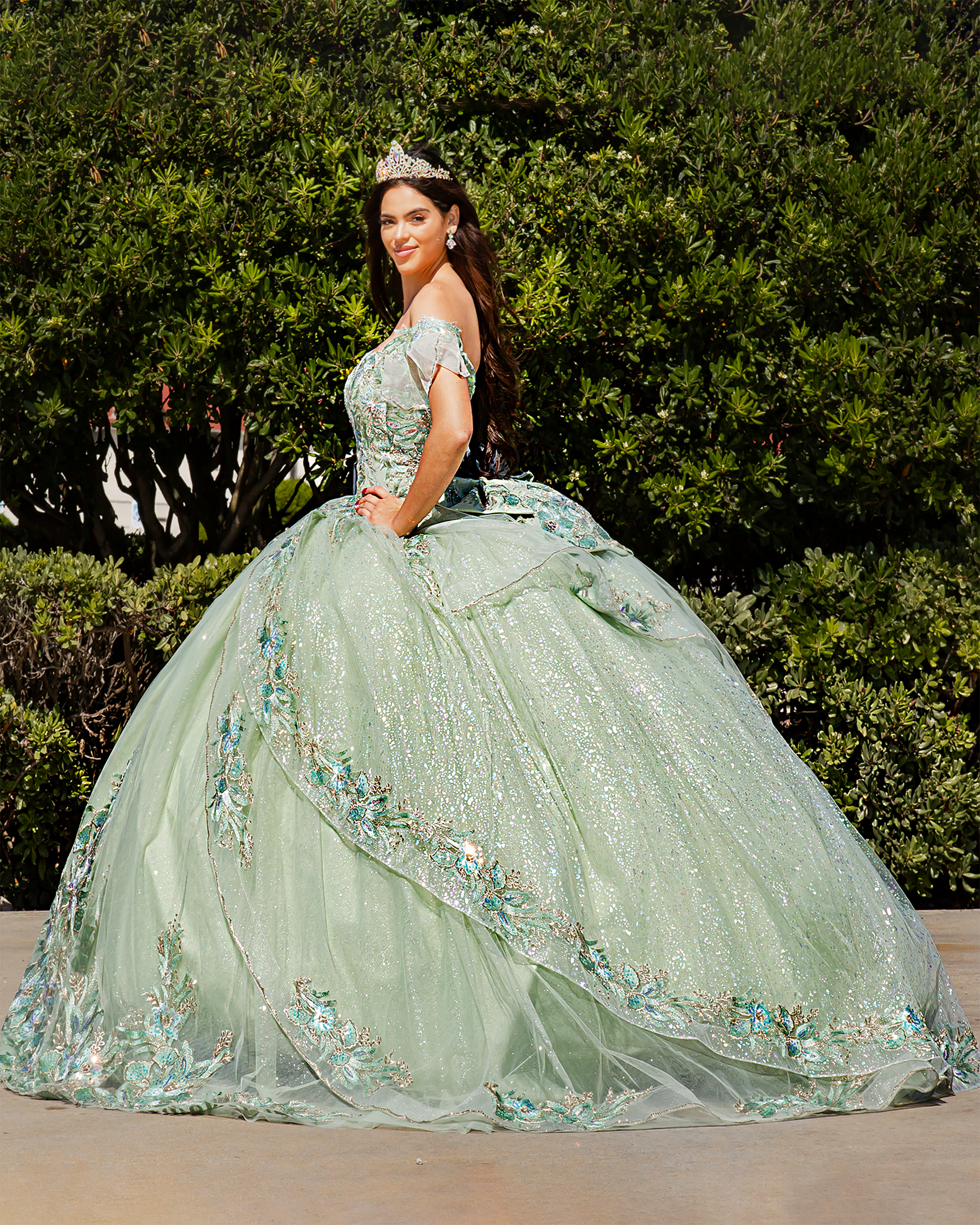Girl in a sage green floral quince dress with floral details standing in front of green bushes.