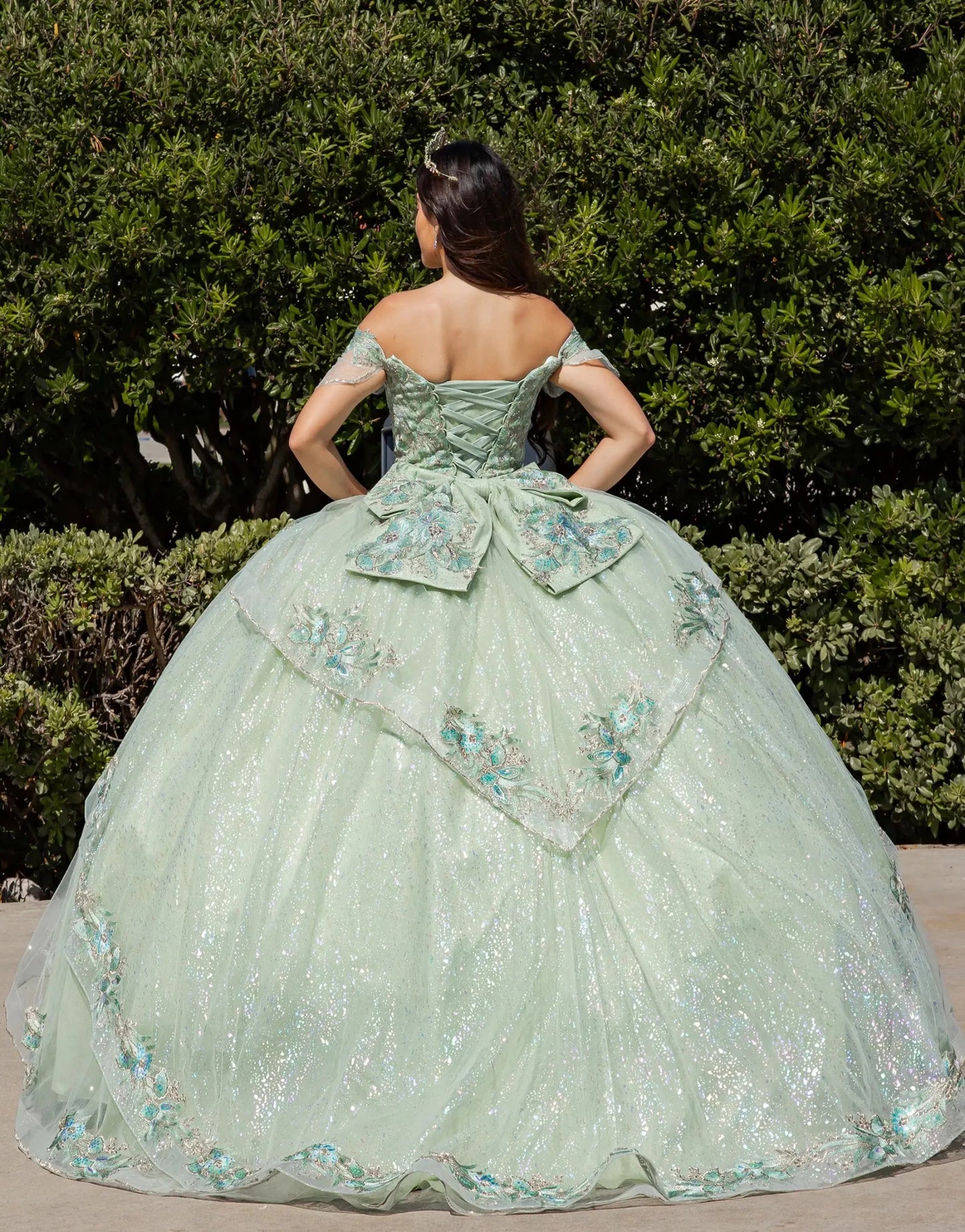 Woman in a light green ball gown with floral embroidery standing in front of green bushes.