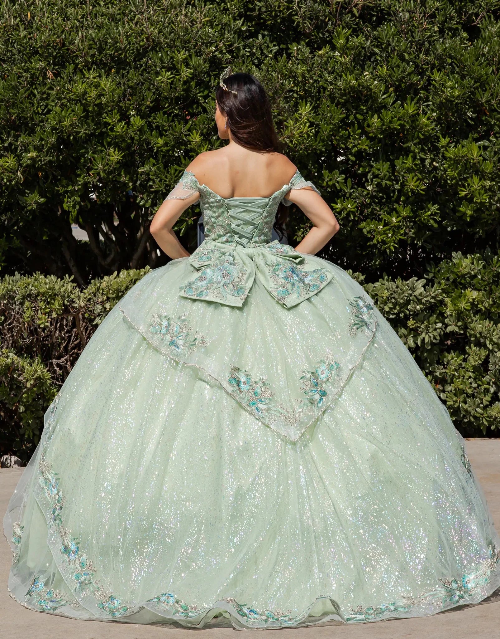 Woman in a light green ball gown with floral embroidery standing in front of green bushes.