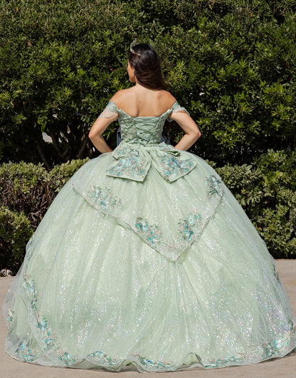 Woman in a light green ball gown with floral embroidery standing in front of green bushes.