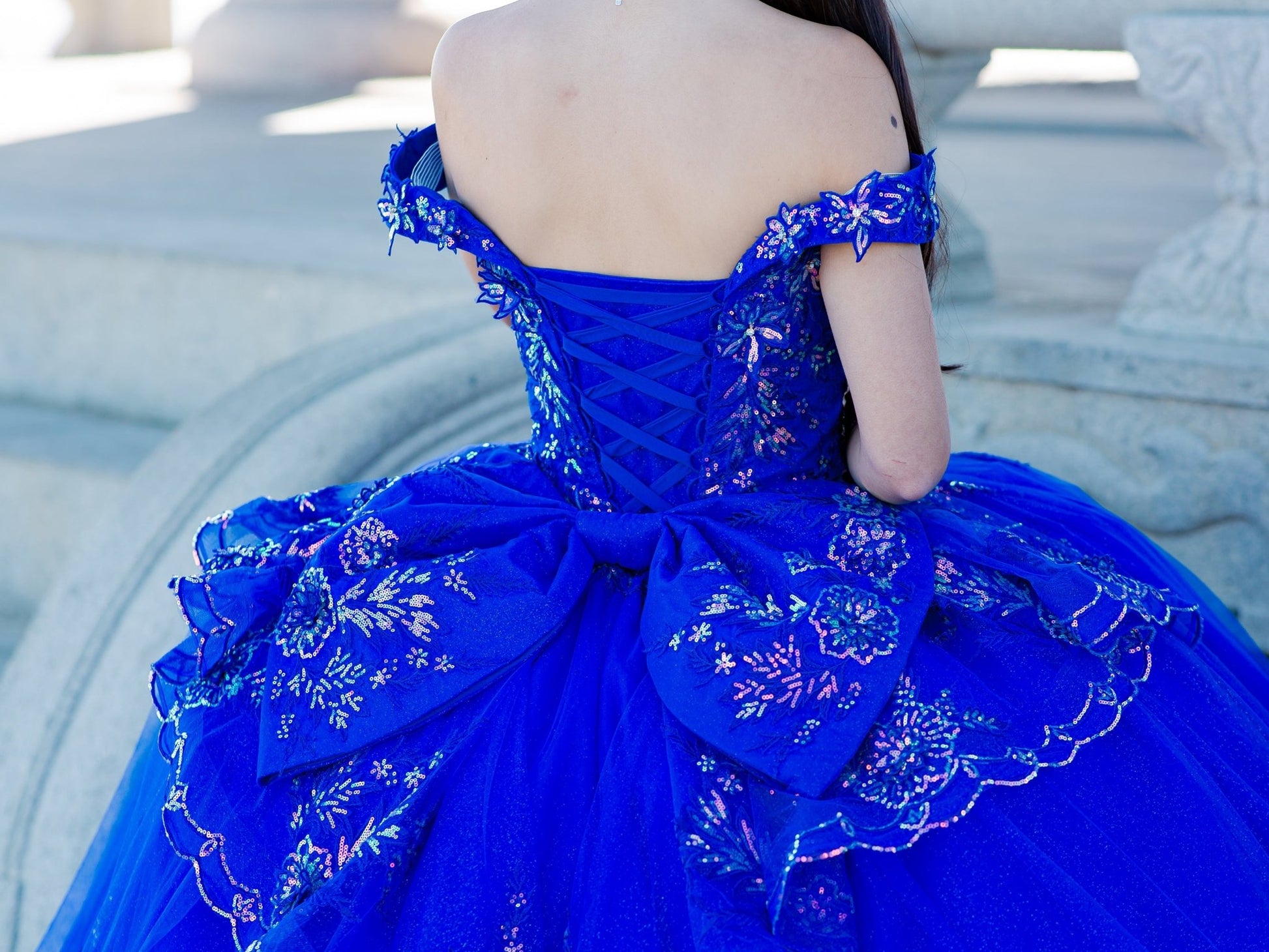 Back zoom view of a woman wearing blue quinceanera dress with beaded applique bodice, lace-up corset, and glitter tulle skirt