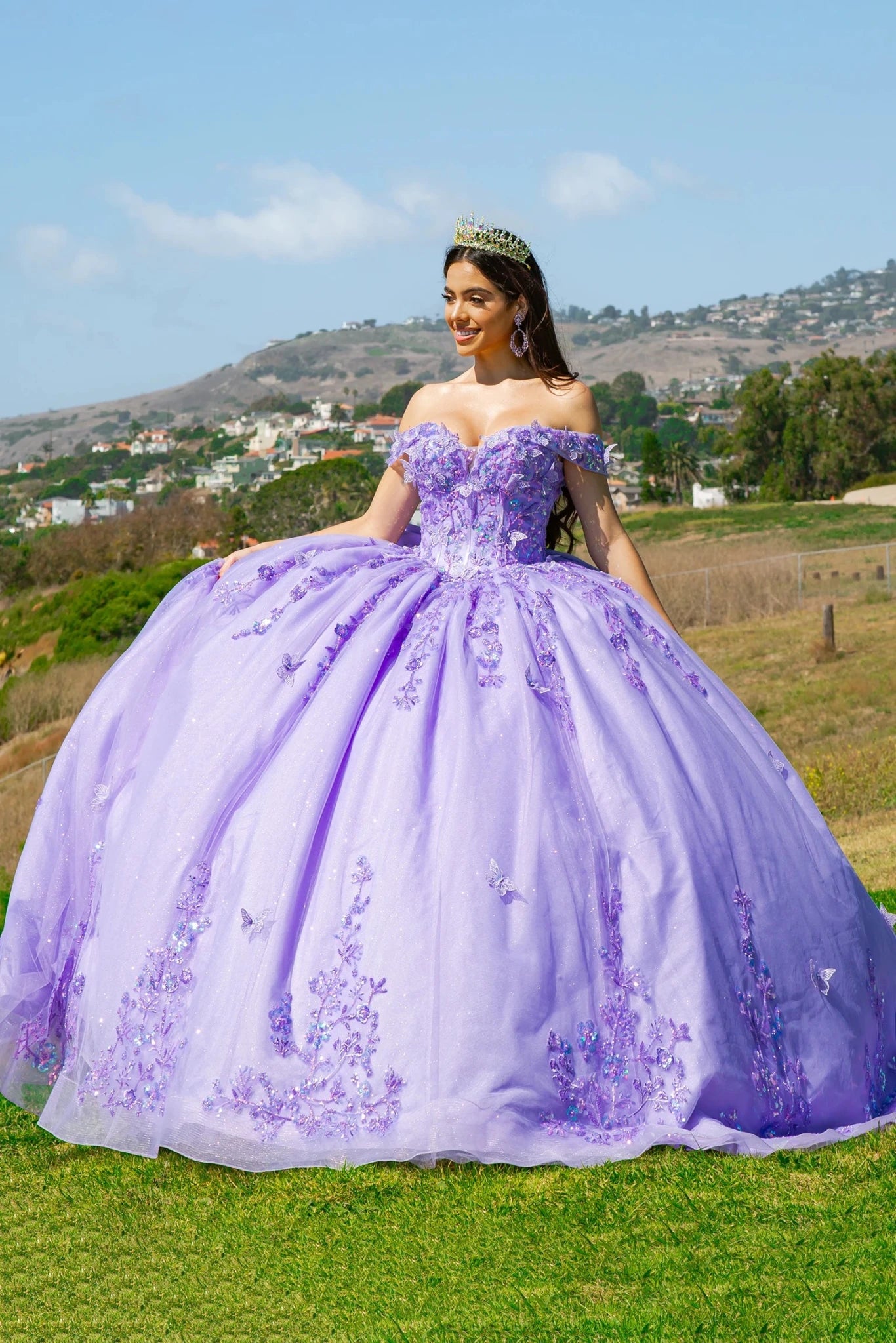 A woman in a lilac quinceañera dress densely adorned with 3D butterfly and floral appliqués, standing outdoors.