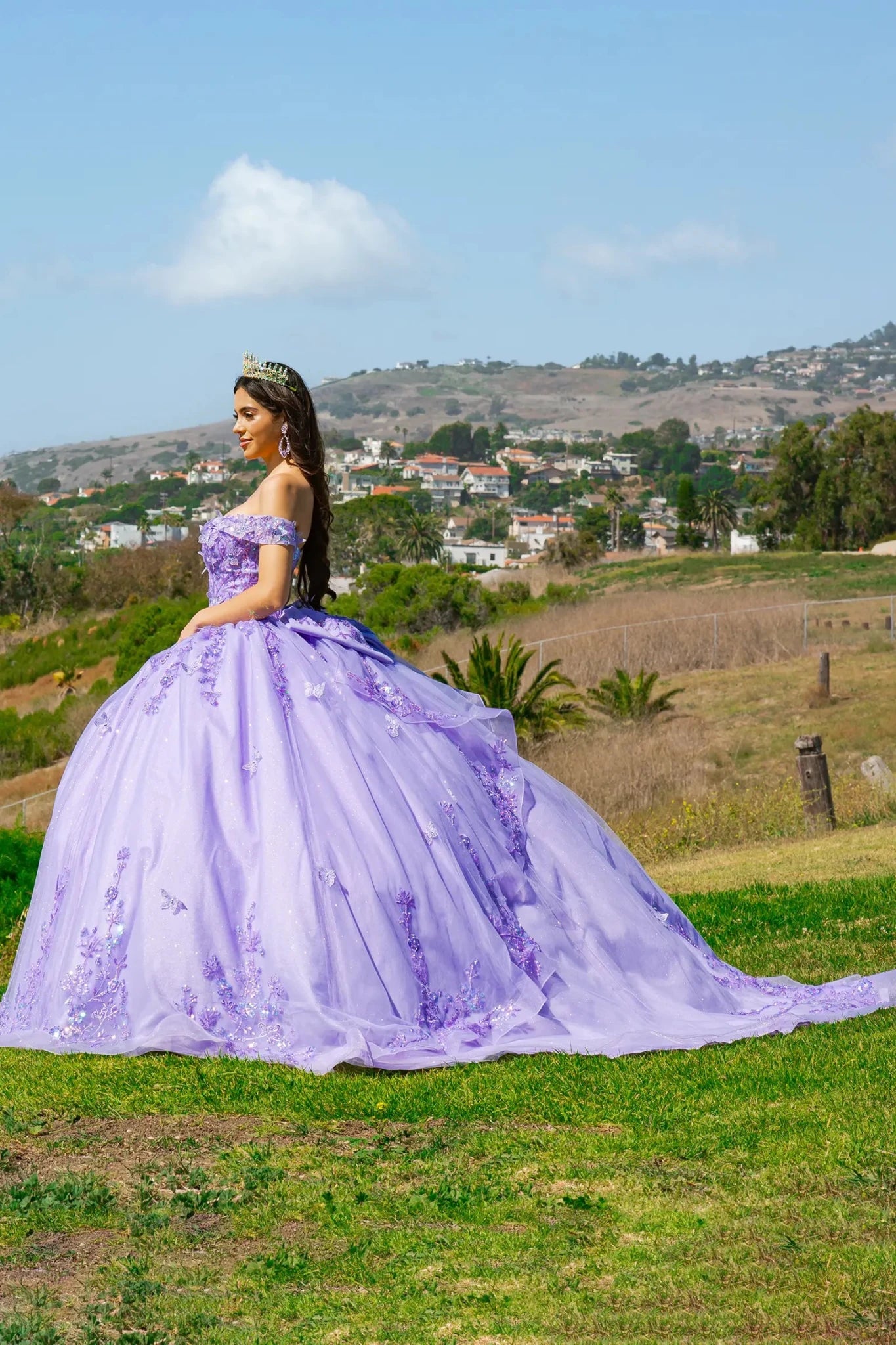 Woman in a lilac ball gown with fluffy skirt standing in a scenic outdoor setting with hills and trees.