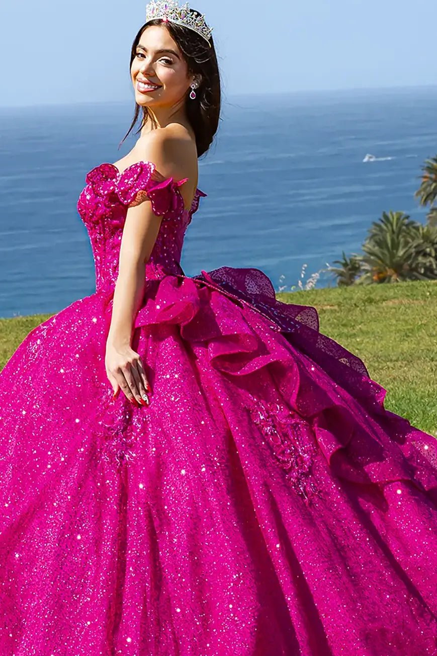 Woman standing side-facing in fuchsia sparkling ball gown with floral bodice embellishments, voluminous skirt, and tiara outdoors on grassy area with ocean and palm trees