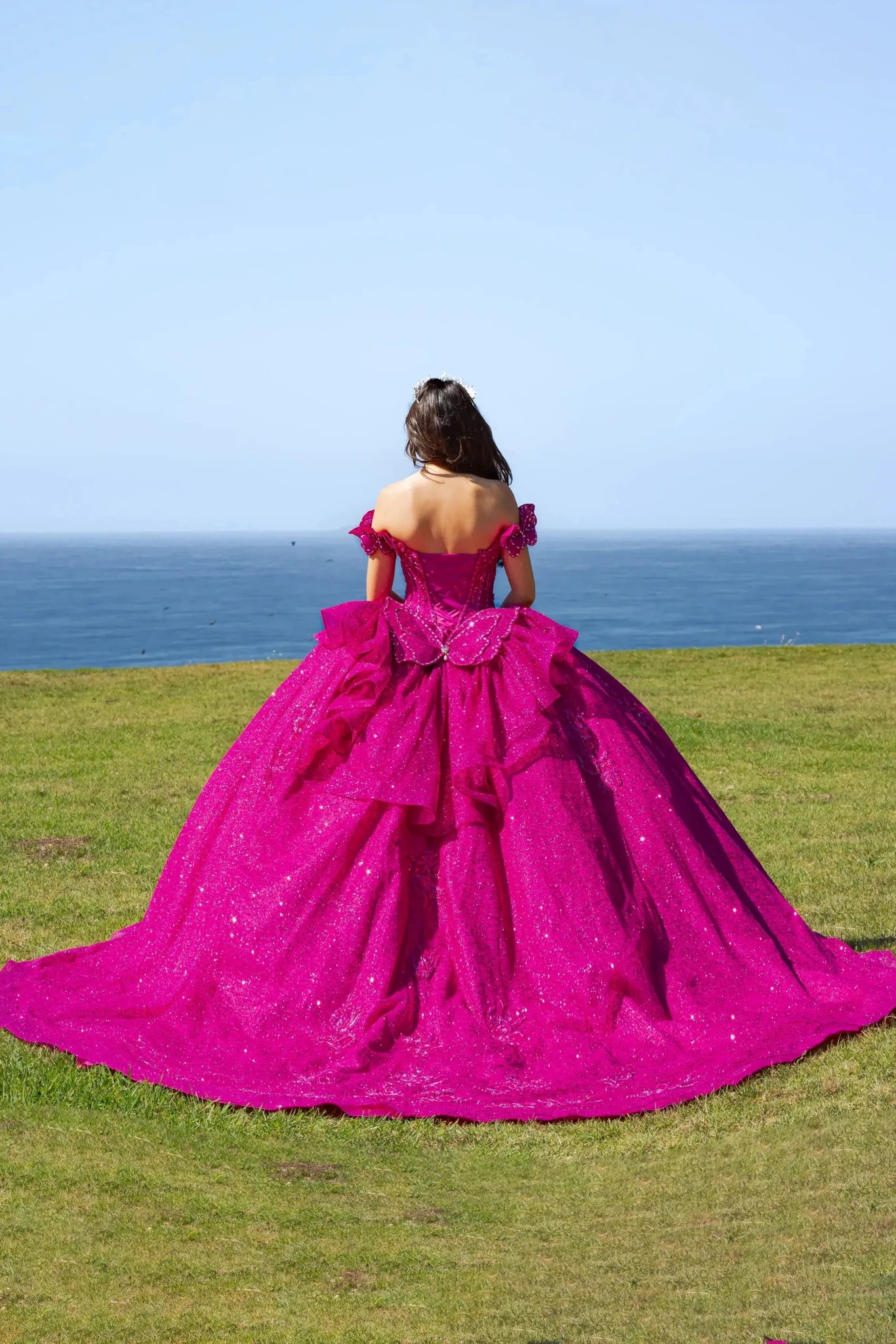 Woman standing back-facing with fuchsia quinceanera ball gown with glittery embellishments, ruffled layers, off-the-shoulder sleeves, and decorative bow outdoors overlooking ocean with clear sky